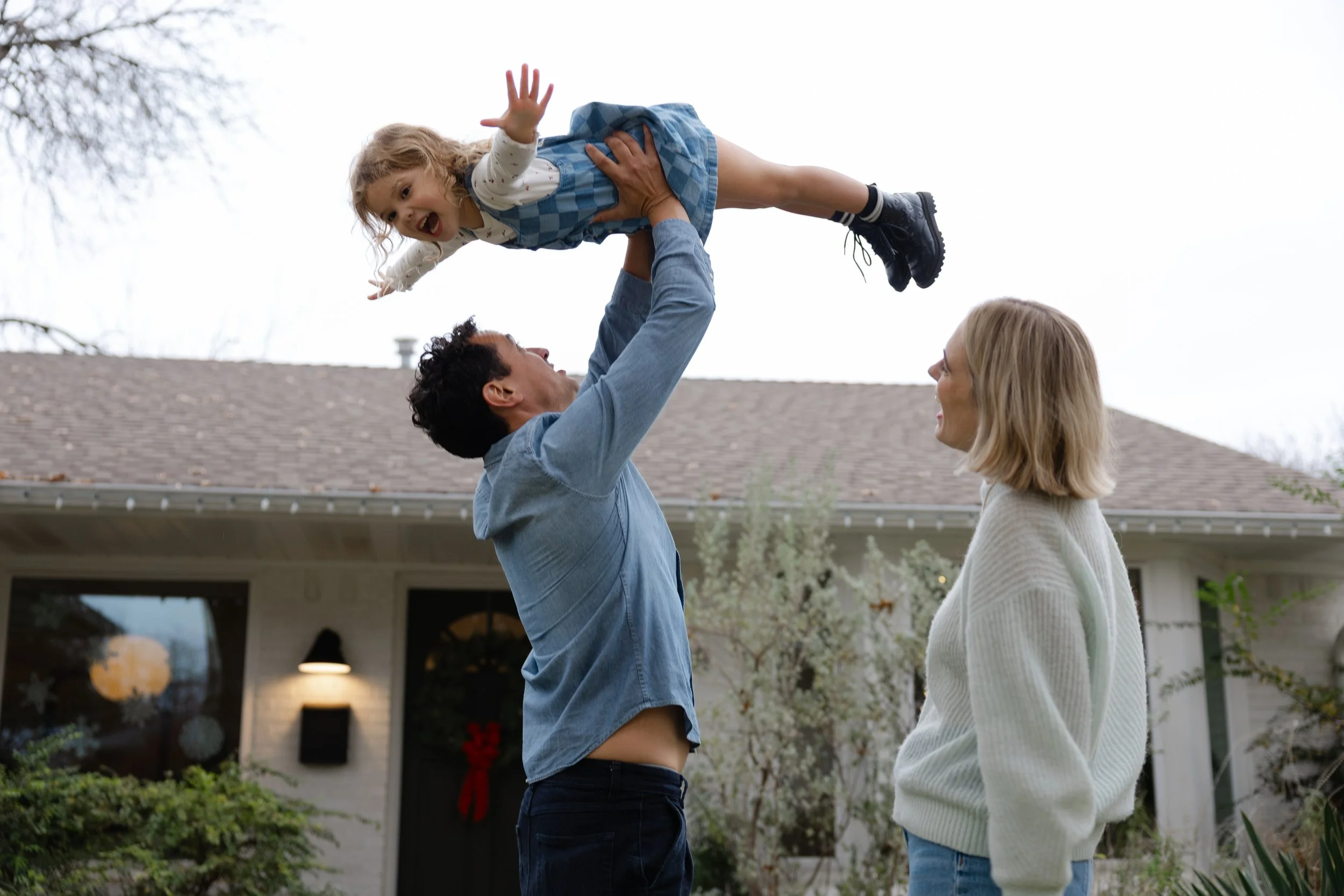 A man lifting a young girl in the air while a woman watches, outside a house with a decorated front door, in a garden with trees and plants.