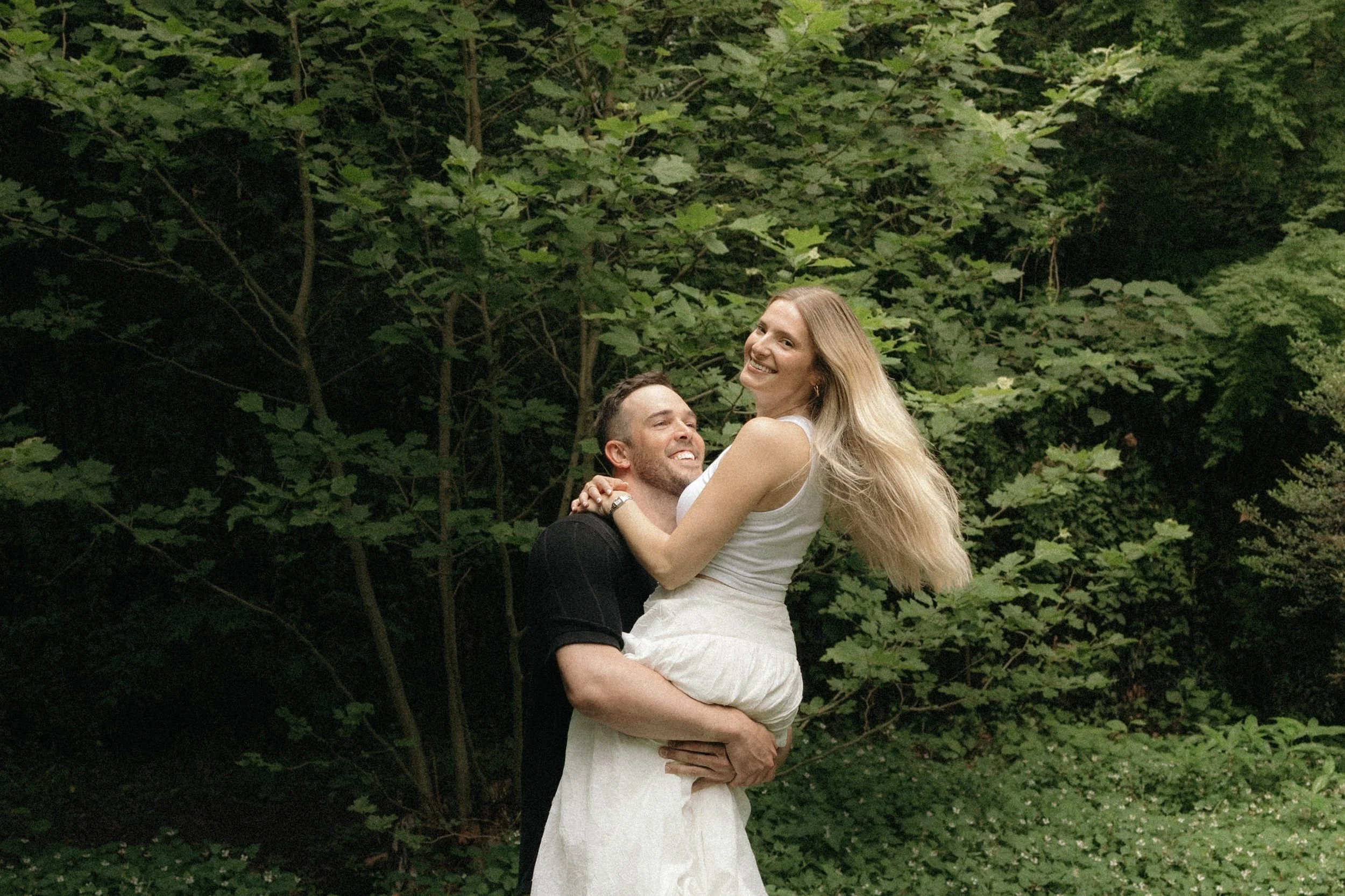A man lifts a woman in a forested area with green trees and foliage, both smiling and looking joyful.
