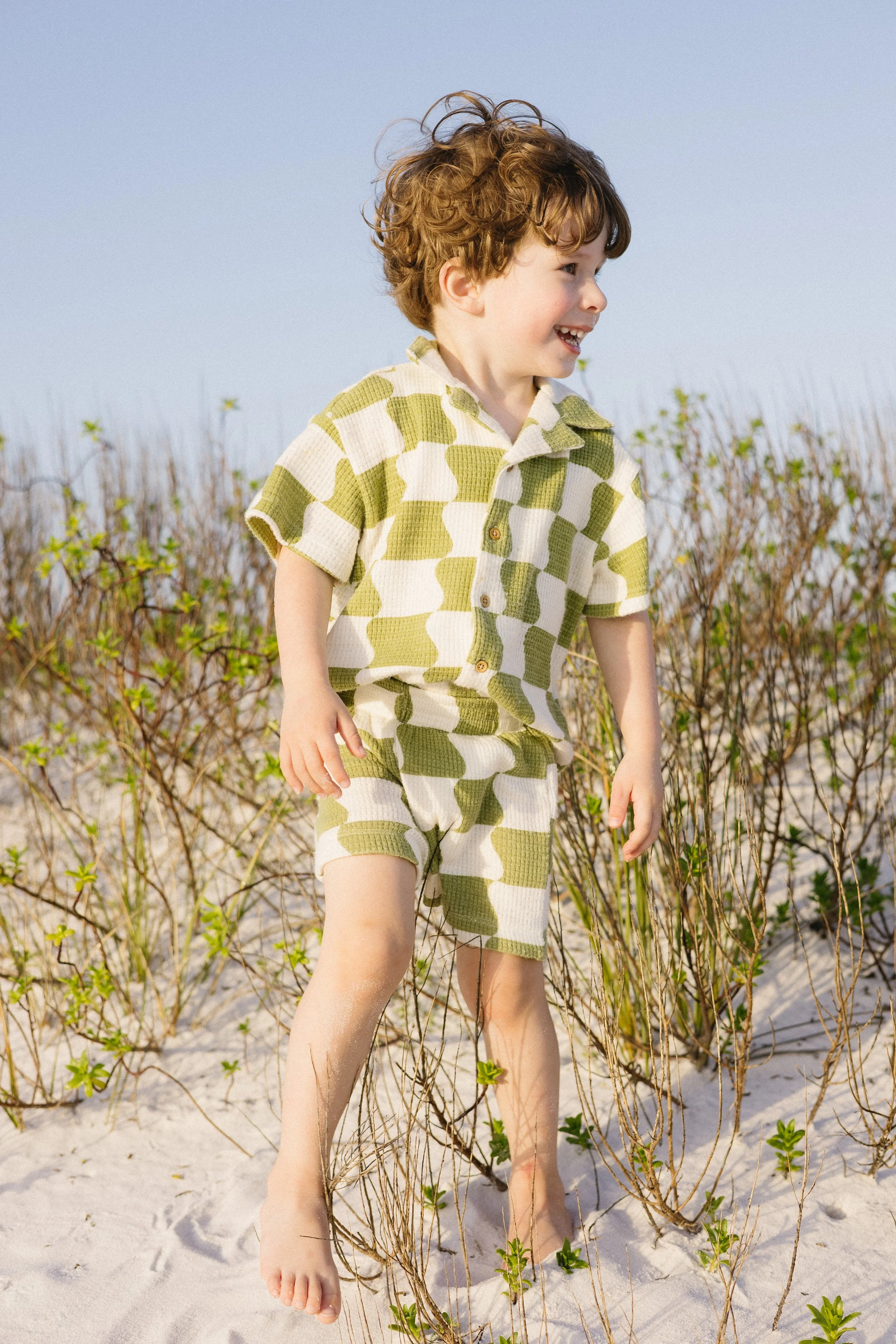 A young boy with curly red hair smiling and wearing a green and white checkered short-sleeved shirt and matching shorts, standing on a sandy beach with low green plants and a clear blue sky in the background.