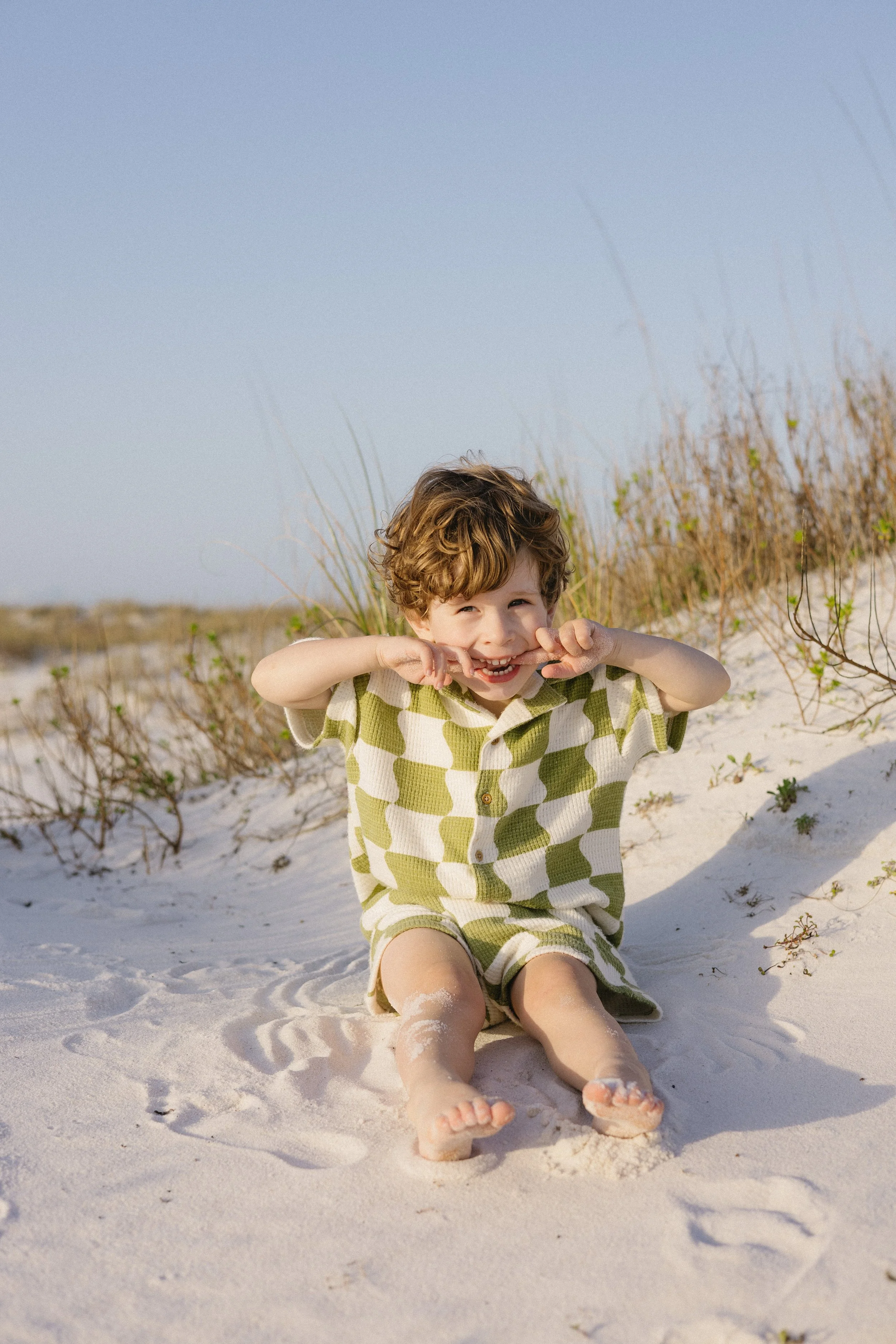 A young boy sitting on the sandy beach, smiling and making a playful gesture with his hands, dressed in a green and white checkered outfit, with the beach grasses in the background.