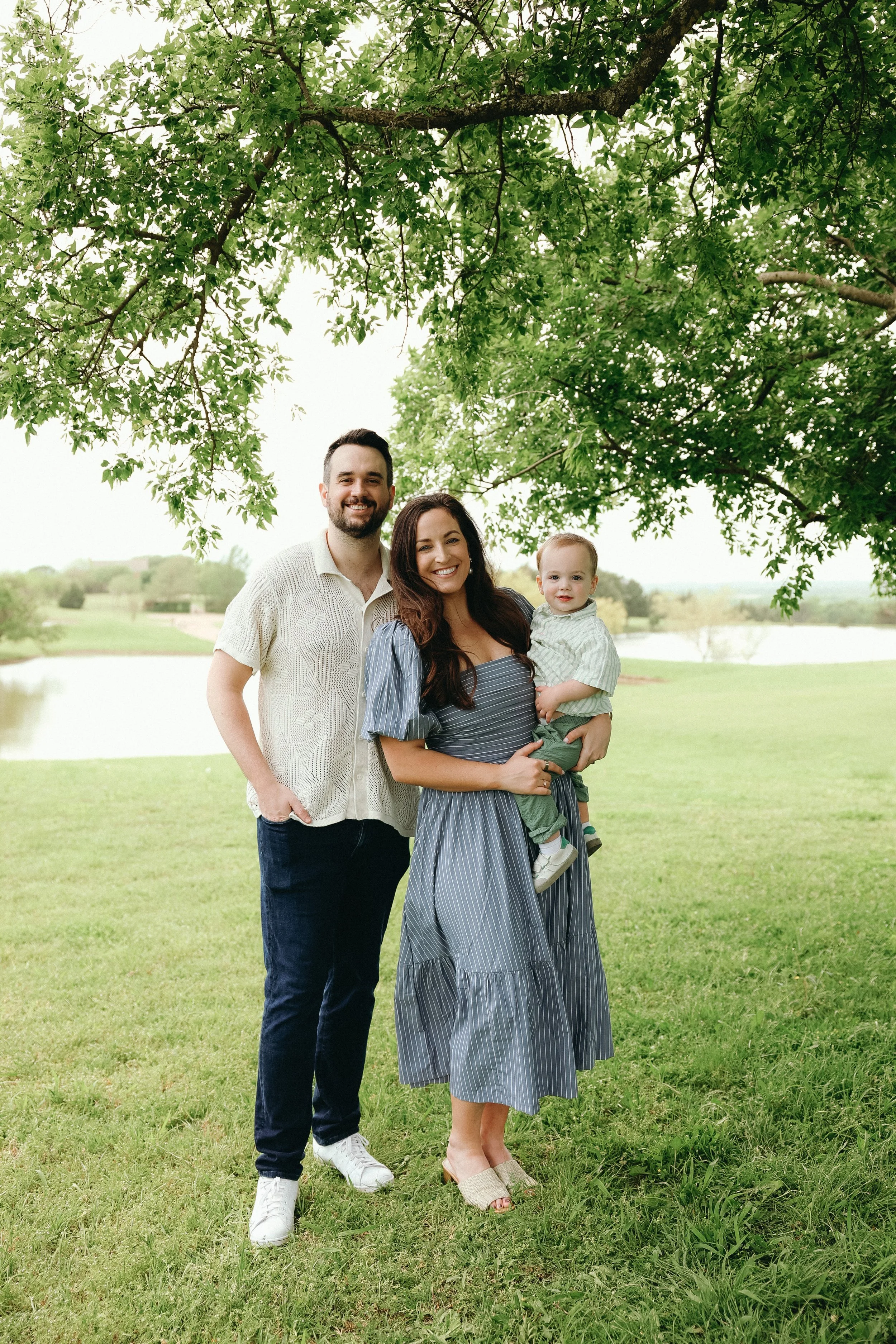A family of three standing outdoors under a large leafy tree, with a lake and green fields in the background, smiling.