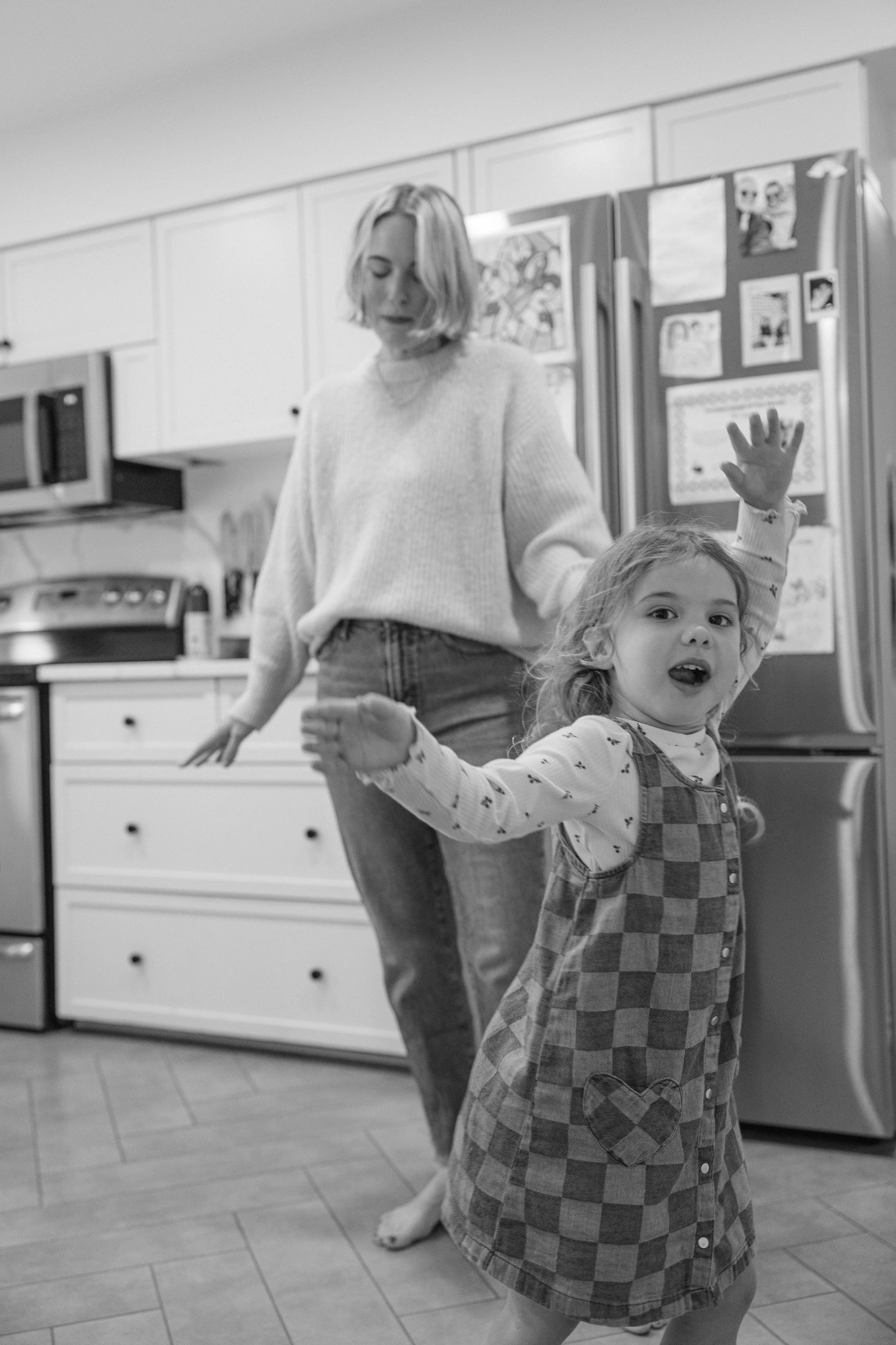 A woman and a young girl dancing and playing in a kitchen, with the girl raising her hand and smiling.
