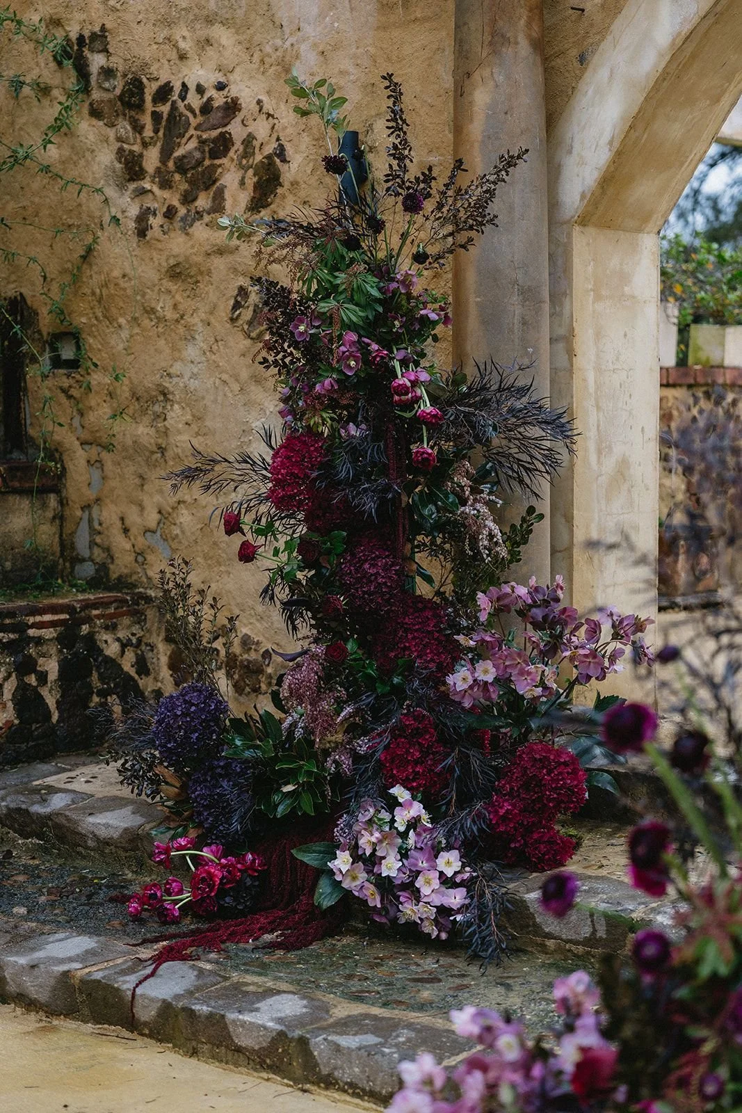 A colorful flower arrangement with pink, purple, and burgundy flowers placed on a stone pathway next to a textured beige wall and stone archway.