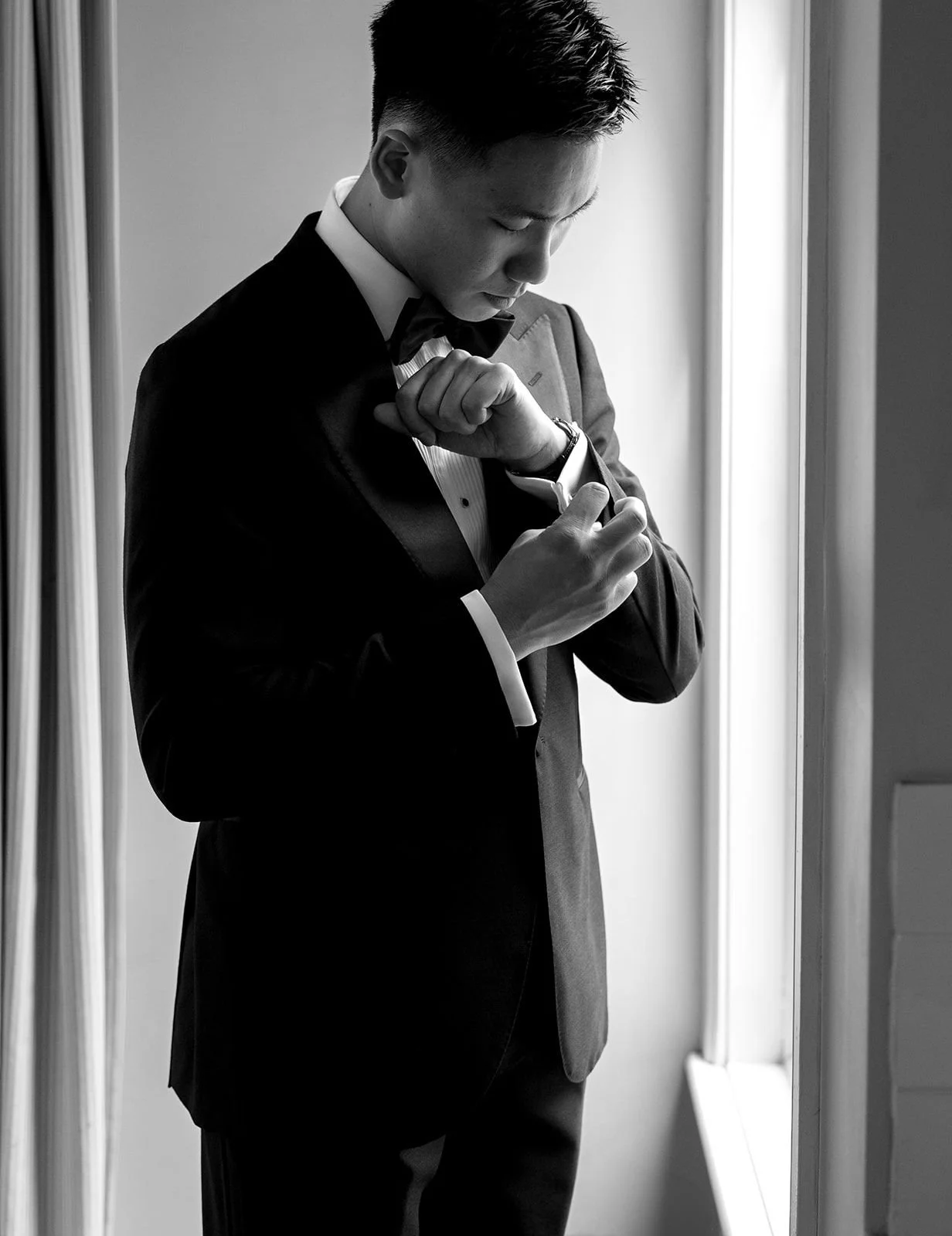 A man in a tuxedo adjusting his cufflinks near a window.