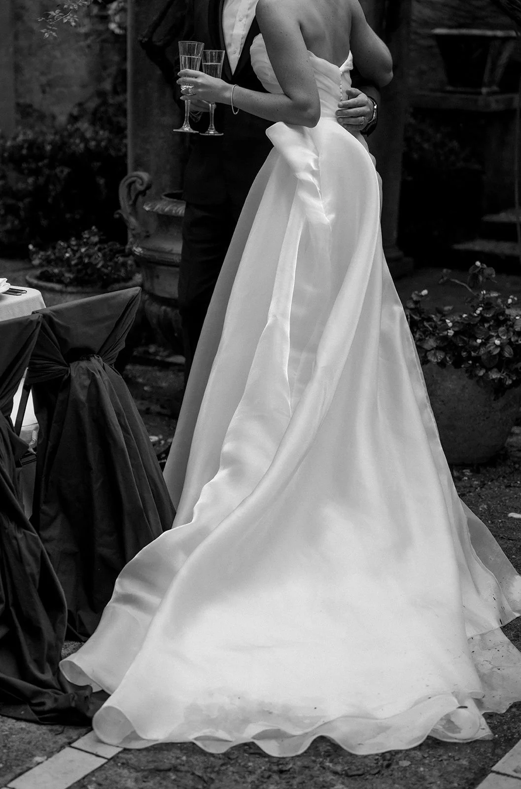 Black and white photo of a bride in a strapless wedding gown with a long train, holding hands with a groom in a suit with a bowtie, both holding champagne glasses, at a wedding reception.