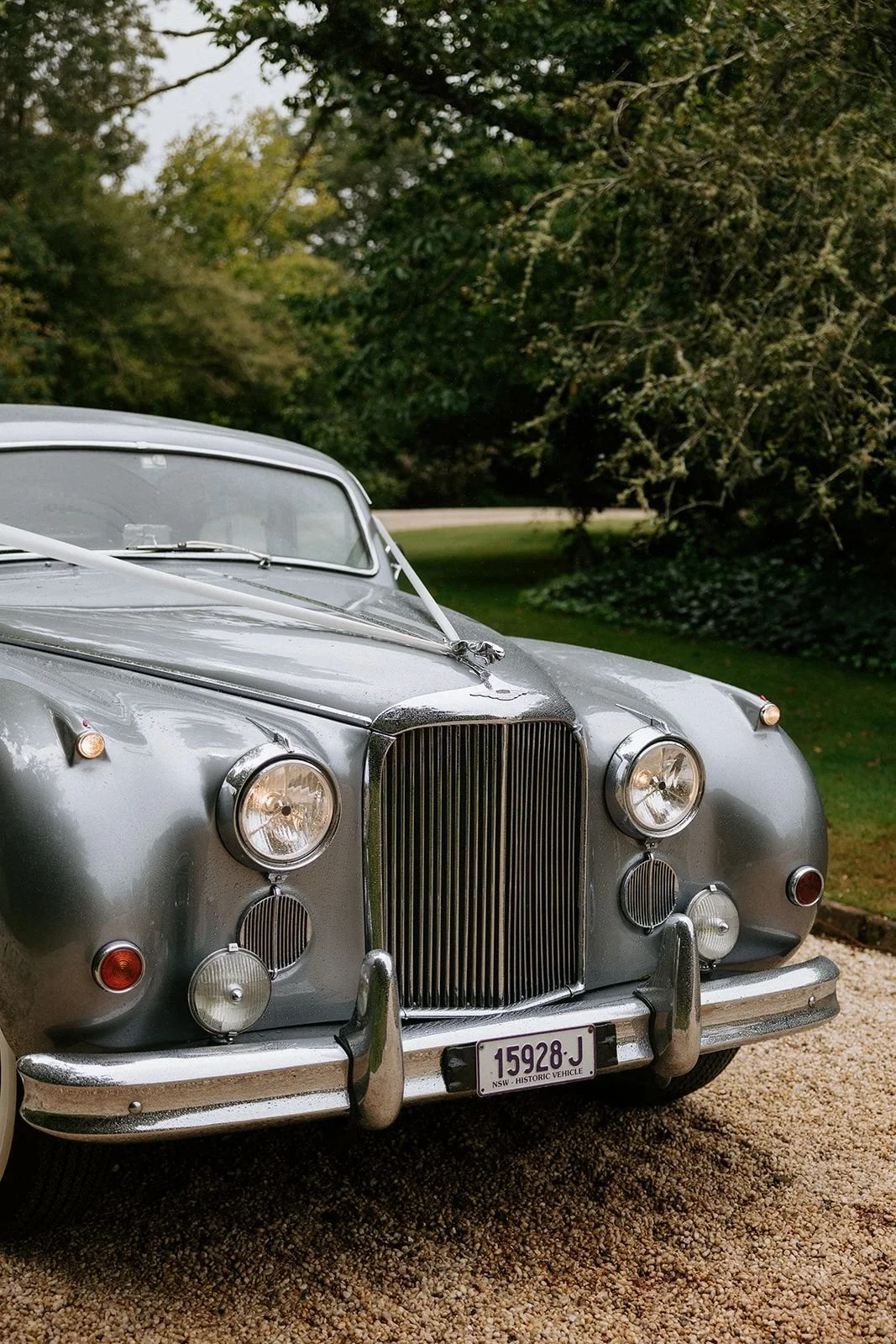 Front view of a vintage silver car with a tall grille, round headlights, and a matte finish, parked on a gravel driveway with green trees in the background.