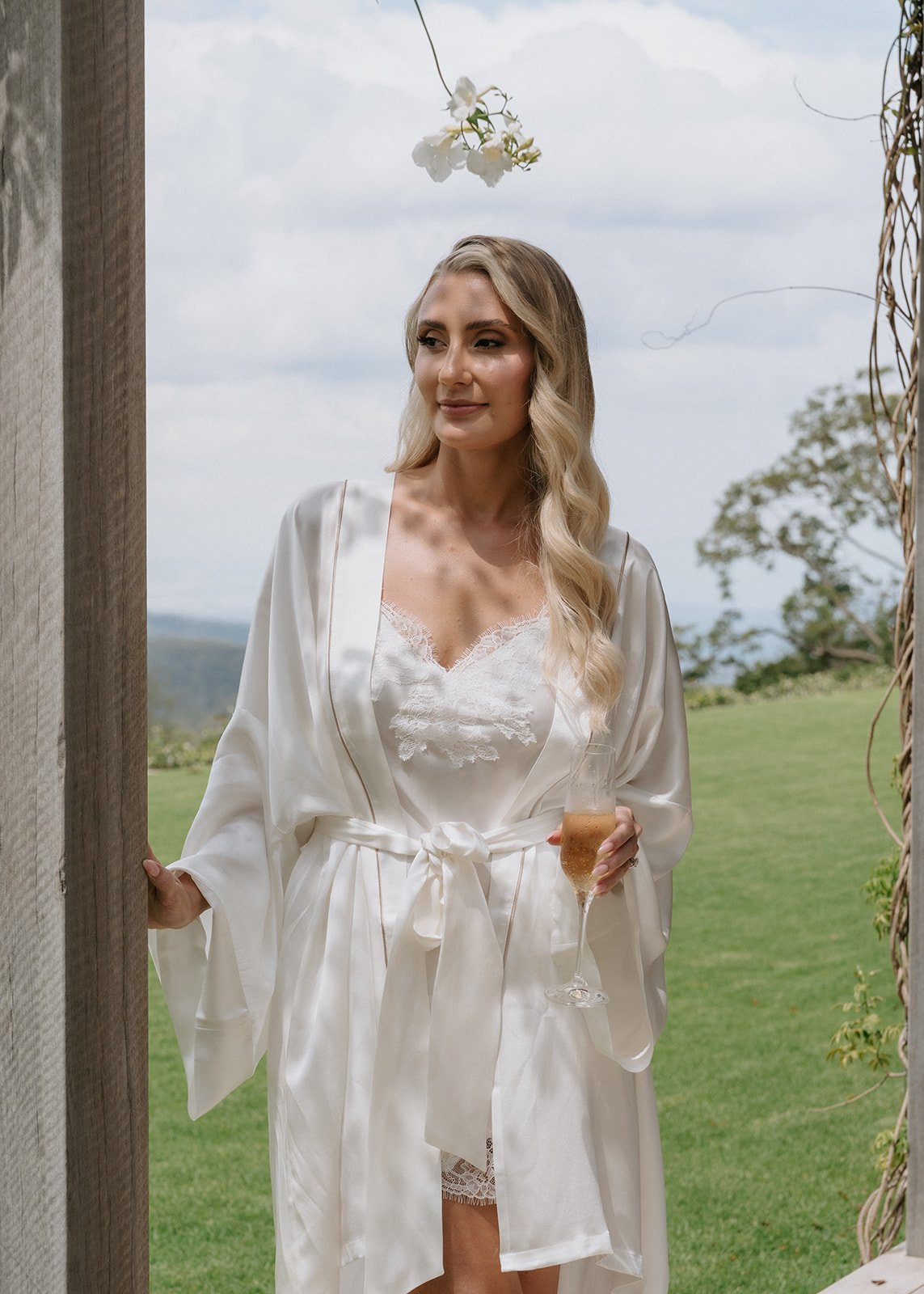 A woman in a white satin robe and lace nightwear holding a glass of champagne, standing outdoors with a lush green background and cloudy sky.