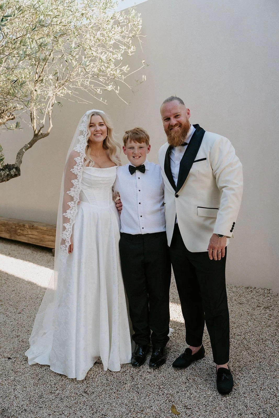 A wedding couple and a young boy posing outdoors. The bride is in a white wedding dress with lace veil, the groom is in a white tuxedo jacket with black lapels, and the boy is in a white dress shirt with black pants and a bow tie.