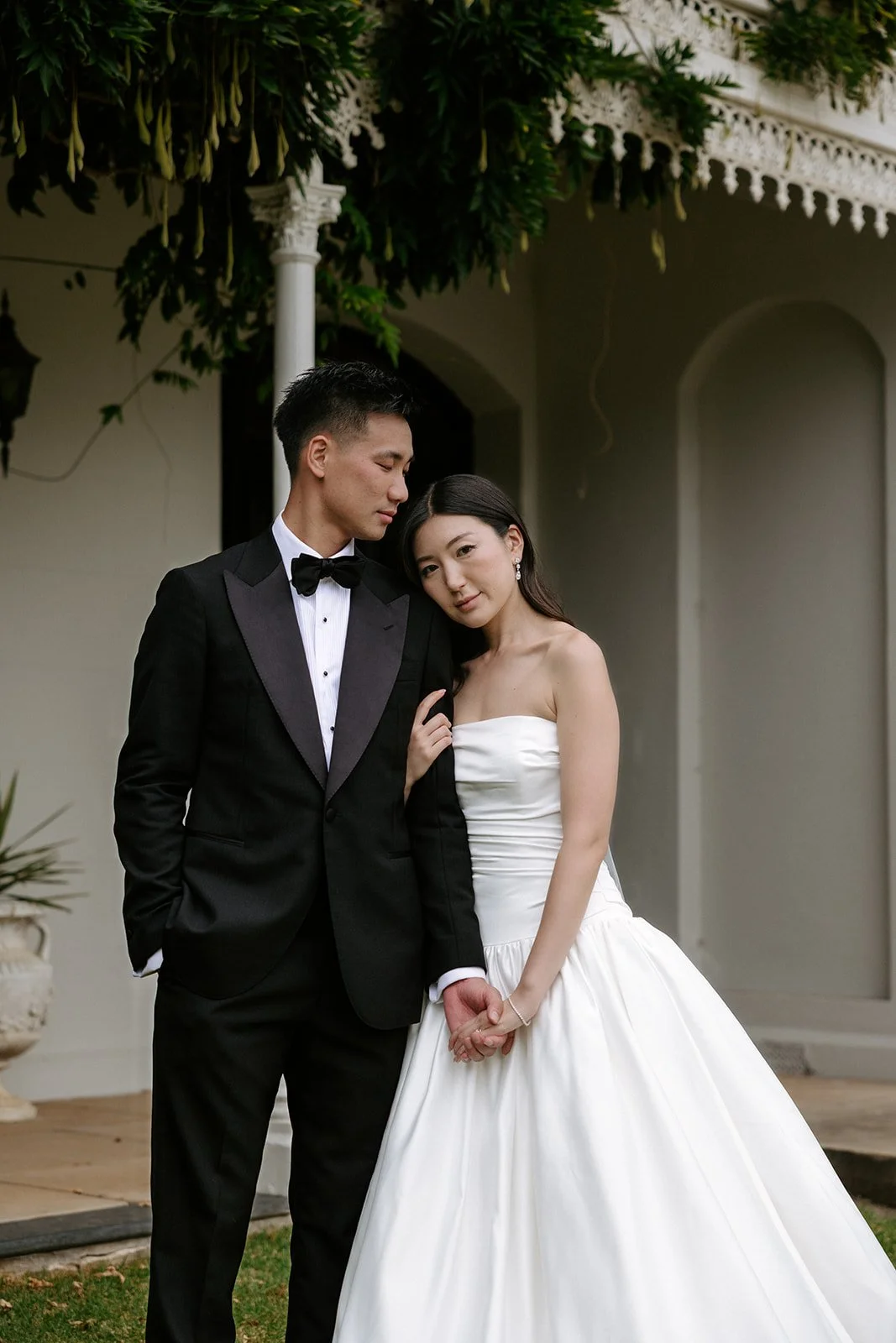 A bride and groom pose in wedding attire outdoors, holding hands, with the bride leaning her head on the groom's shoulder.