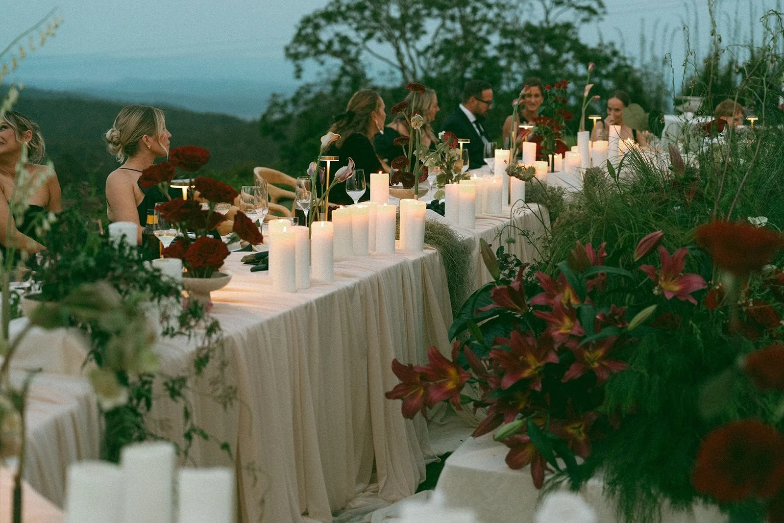 People dressed formally sitting at a long outdoor dinner table with candles and floral arrangements during sunset against a backdrop of trees and mountains.