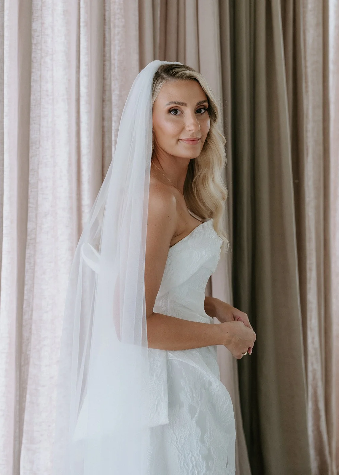 A bride in a white wedding dress and veil standing in front of beige curtains, looking at the camera.
