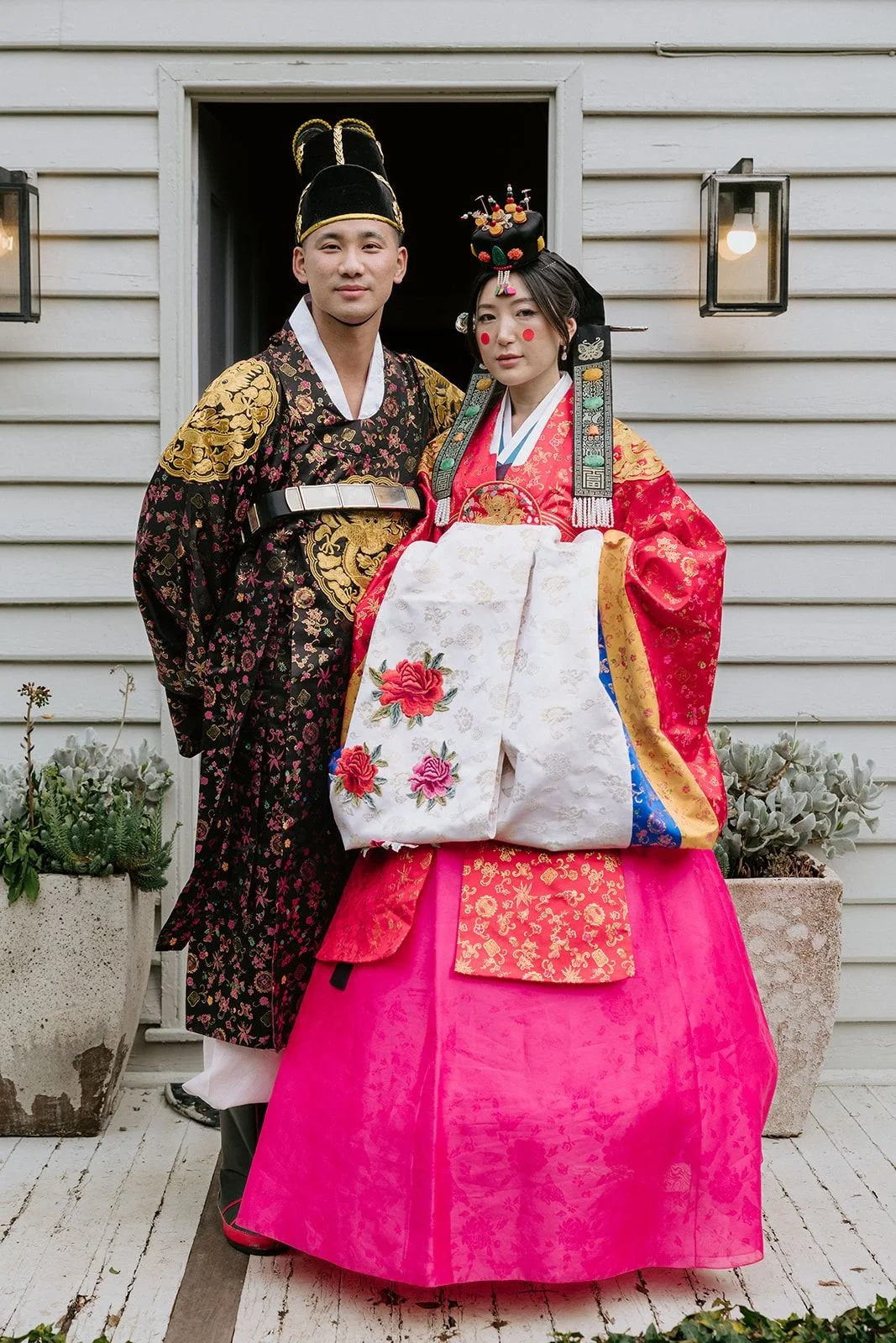 A man and woman dressed in traditional Korean wedding hanbok, standing outside a house with a white wooden wall, potted plants, and a lantern. The man wears a black and gold patterned jacket with a black hat, and the woman wears a vibrant red and pin