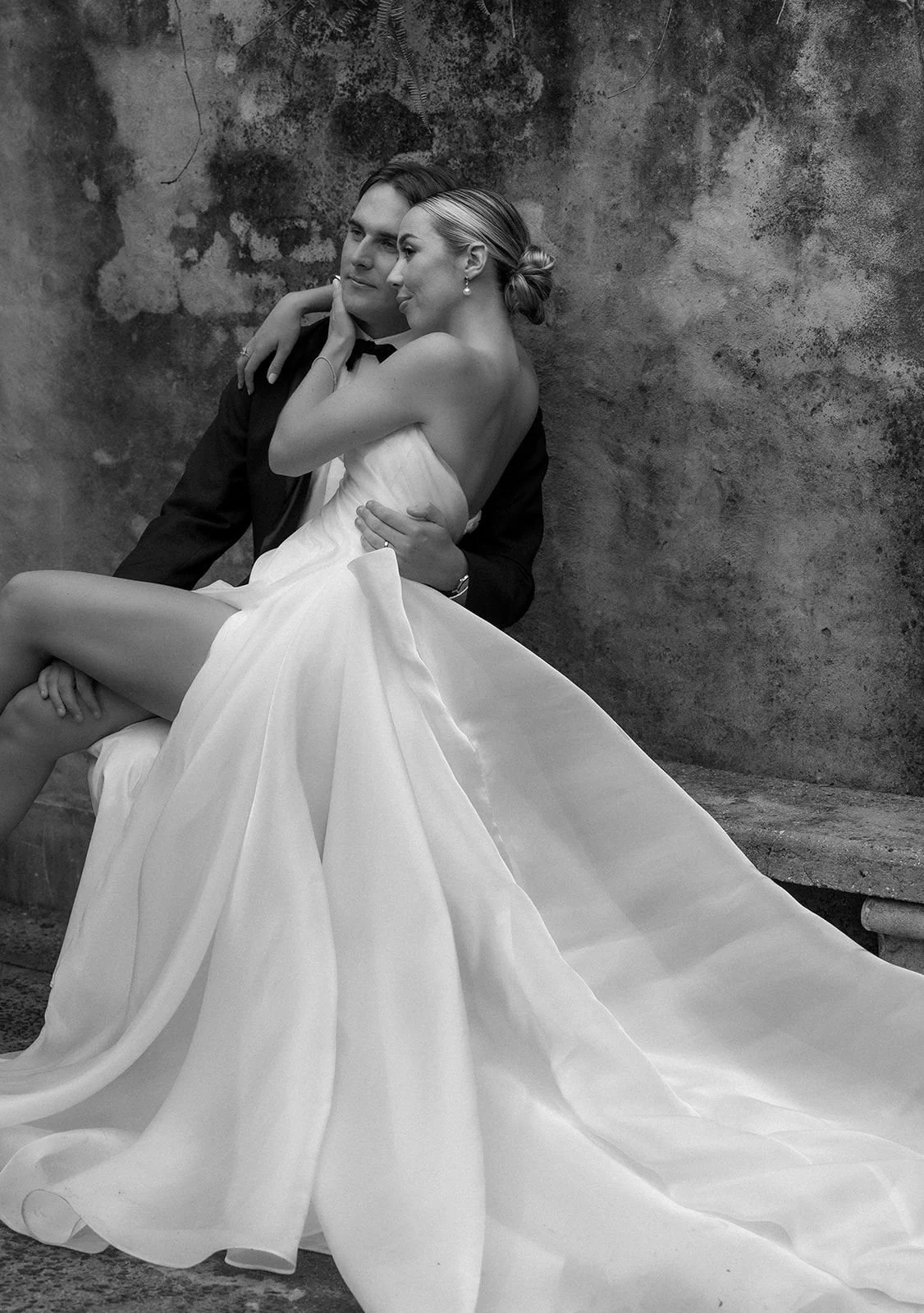 A bride and groom sitting together against a textured wall, with the bride wearing a strapless wedding dress and the groom in a suit, in a romantic pose.