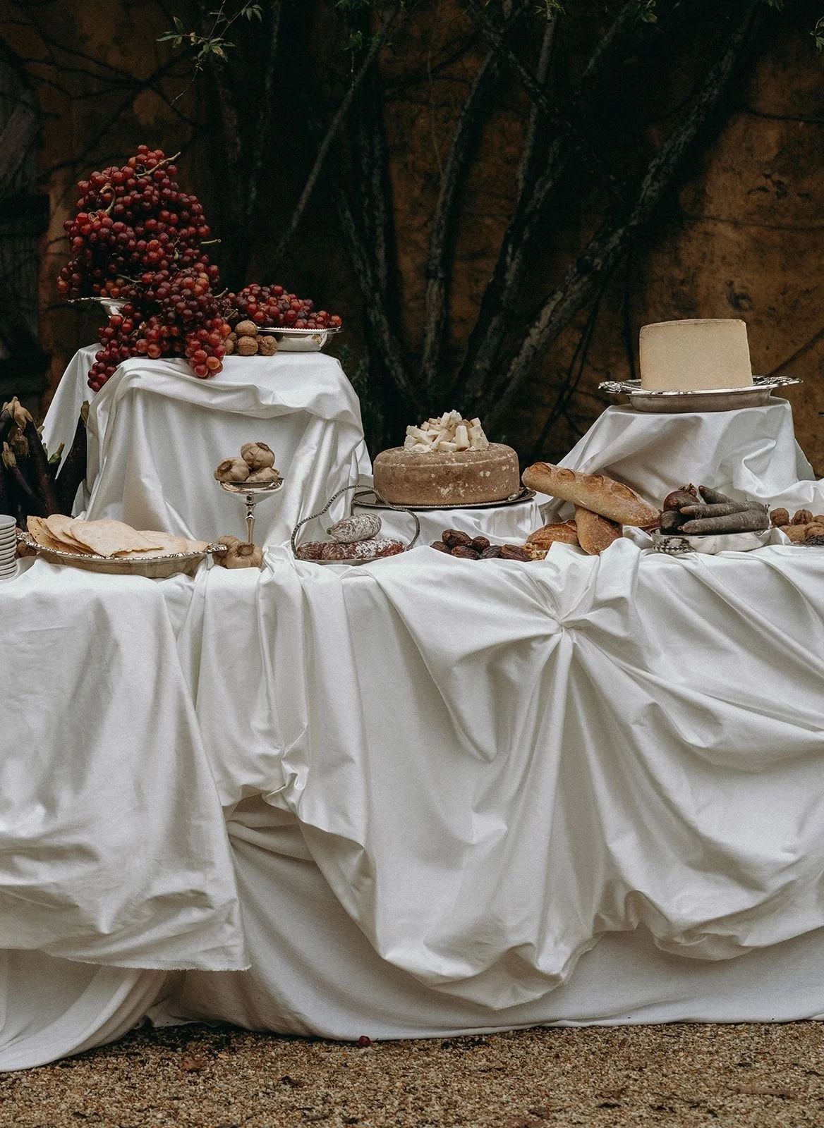 A rustic outdoor cheese and bread display with grapes, nuts, and various cheeses on a table draped in white fabric.