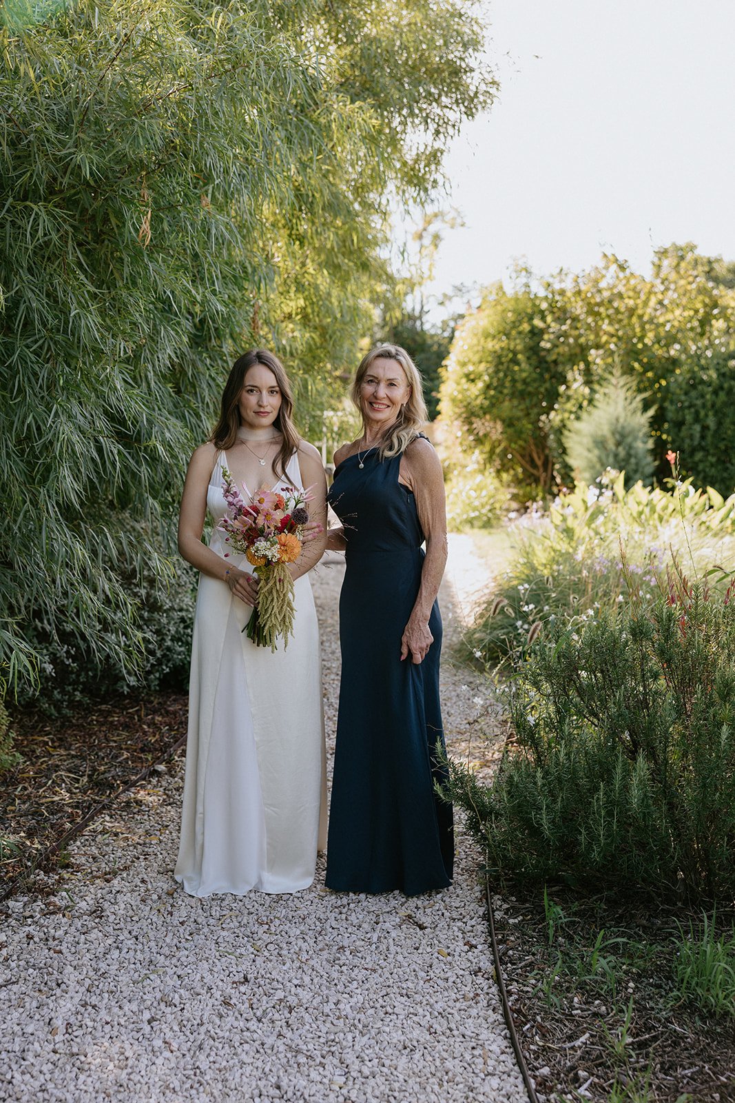A young woman in a white dress holding a colorful bouquet standing next to an older woman in a dark blue dress on a gravel pathway surrounded by lush greenery and trees.