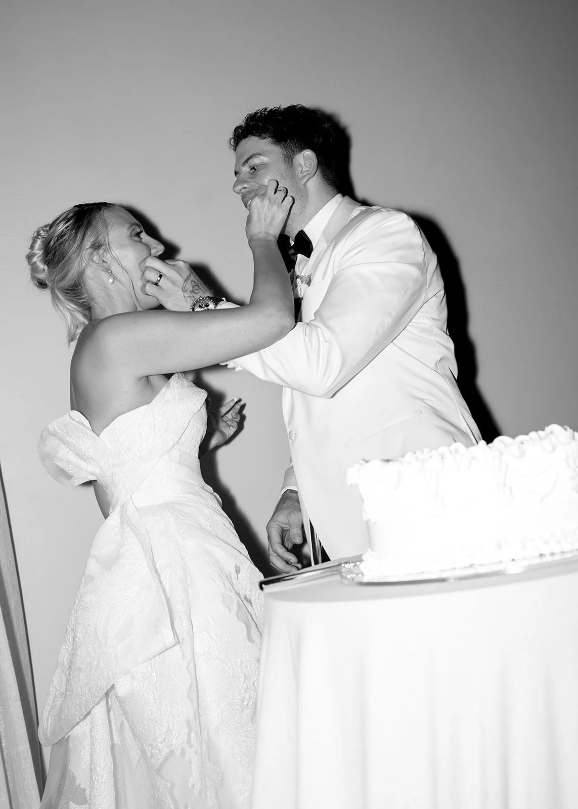 Black and white photo of a bride and groom at their wedding, standing close together, smiling, with a wedding cake nearby.