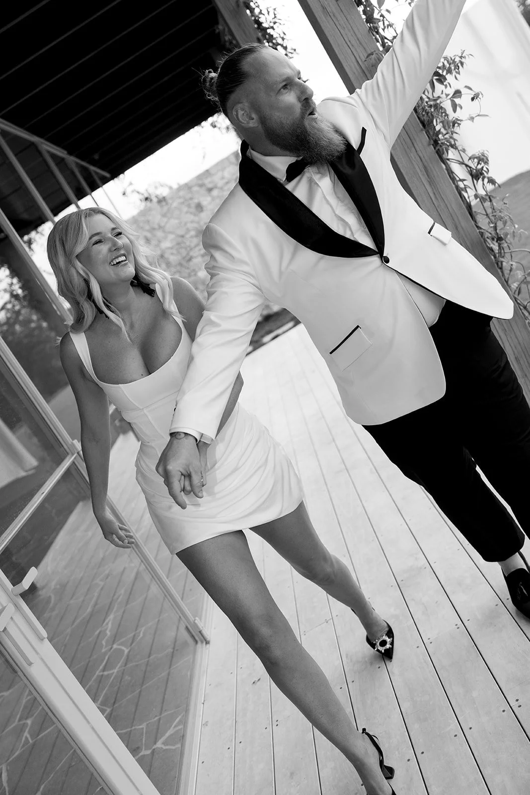 A black-and-white photo of a man and woman holding hands and dancing at a celebration or event. The man has a beard, long hair tied back, and is wearing a white tuxedo jacket with black accents. The woman has shoulder-length blonde hair, is wearing a