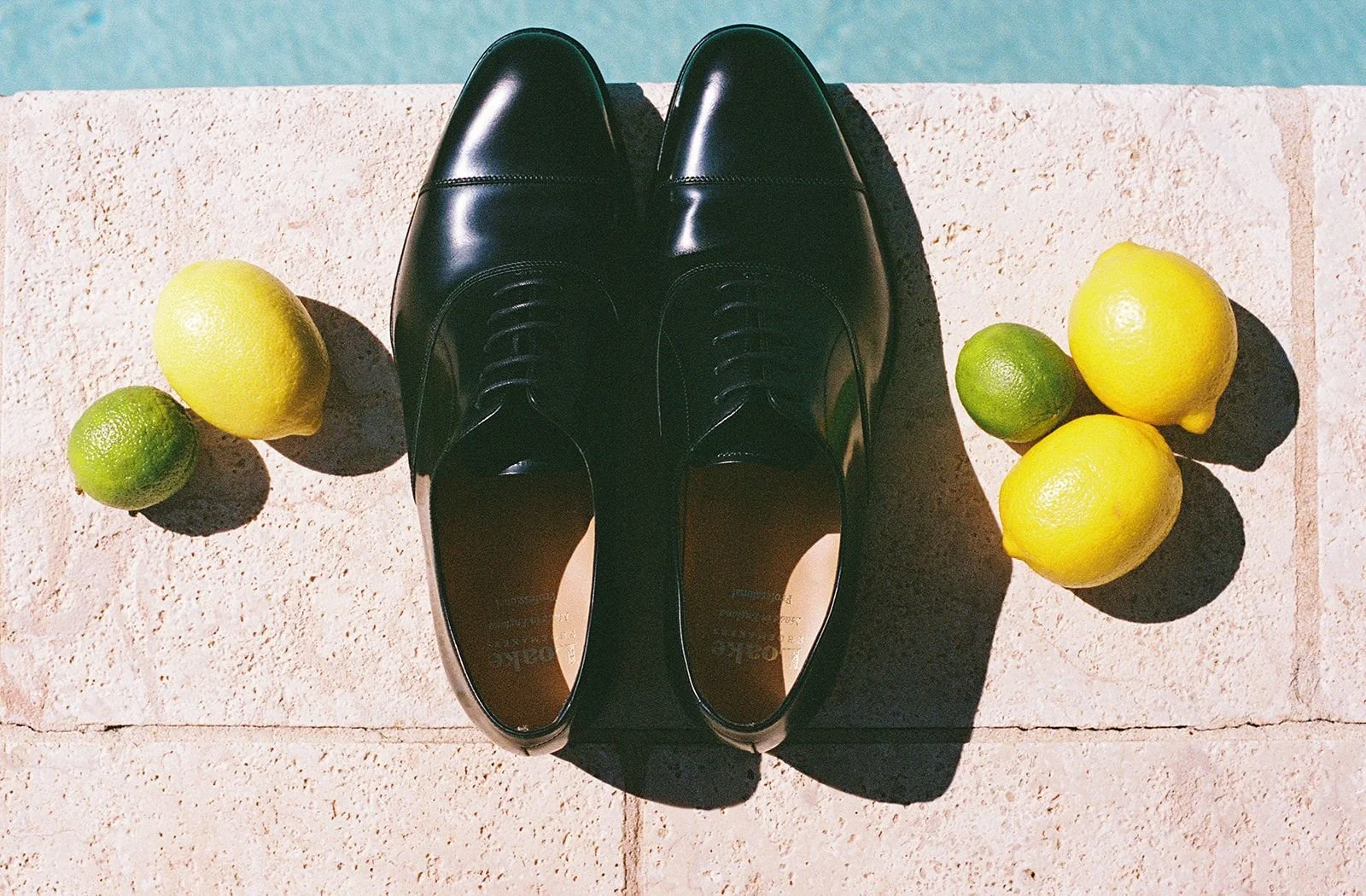 A pair of shiny black dress shoes placed on a stone ledge, with three lemons and two limes arranged around them, casting shadows in sunlight.