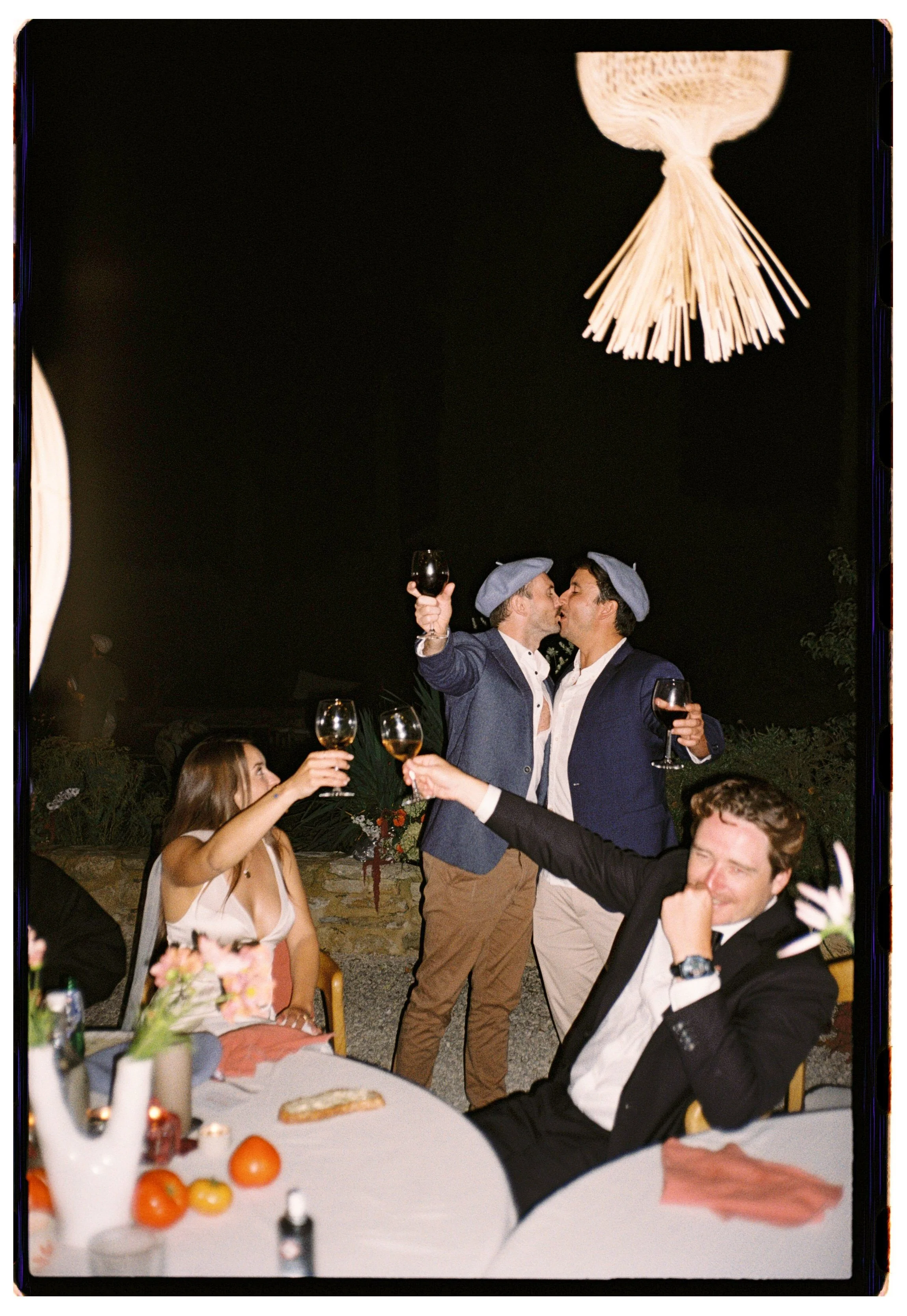 Group of friends celebrating at night, two men in blue suits and gray berets kiss while holding wine glasses, others toast with drinks, a woman in a white dress watches, and a man in a tuxedo laughs at a decorated table with oranges, flowers, and can