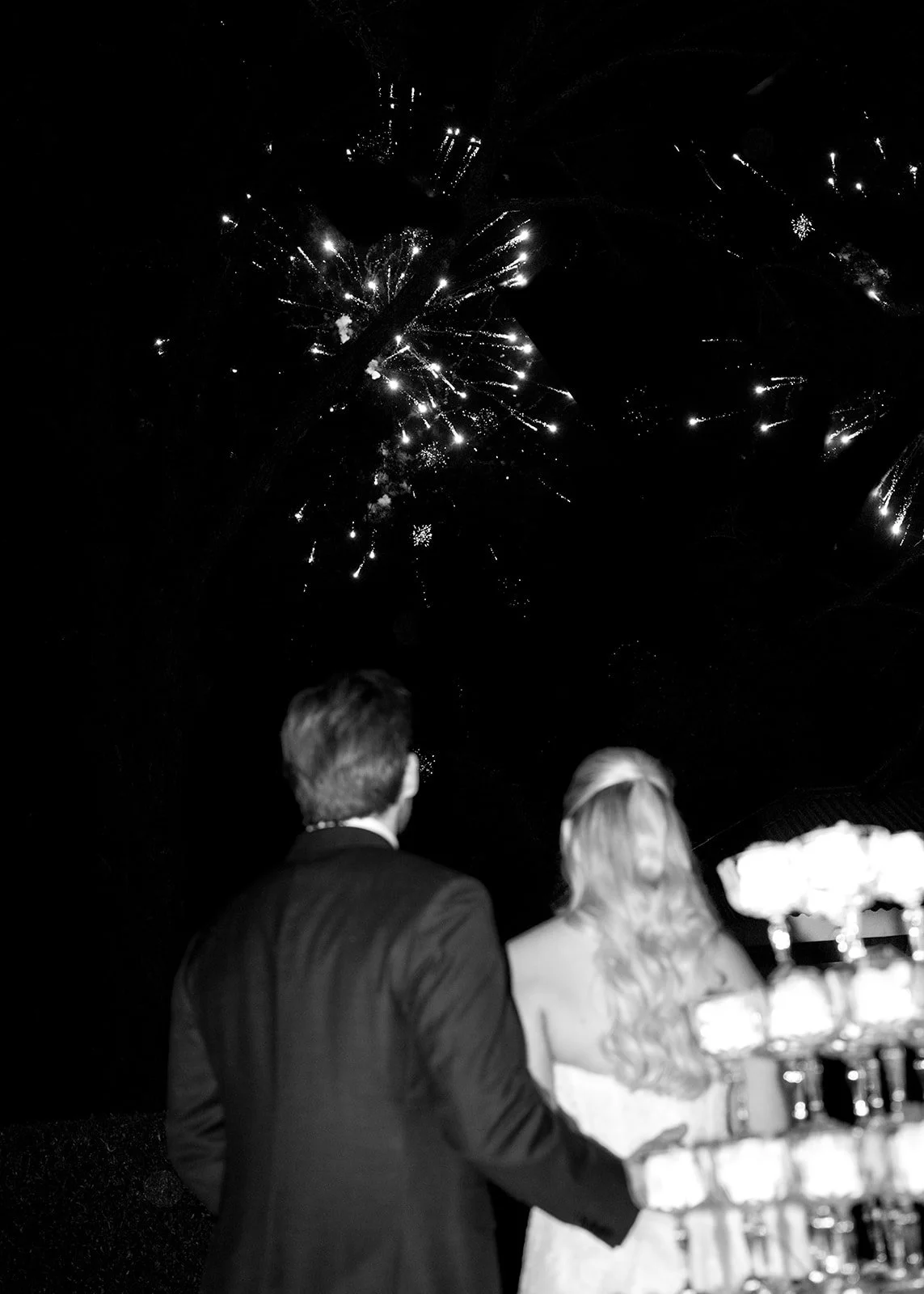 A couple in formal attire watching fireworks in the night sky during what appears to be a celebration or wedding reception.