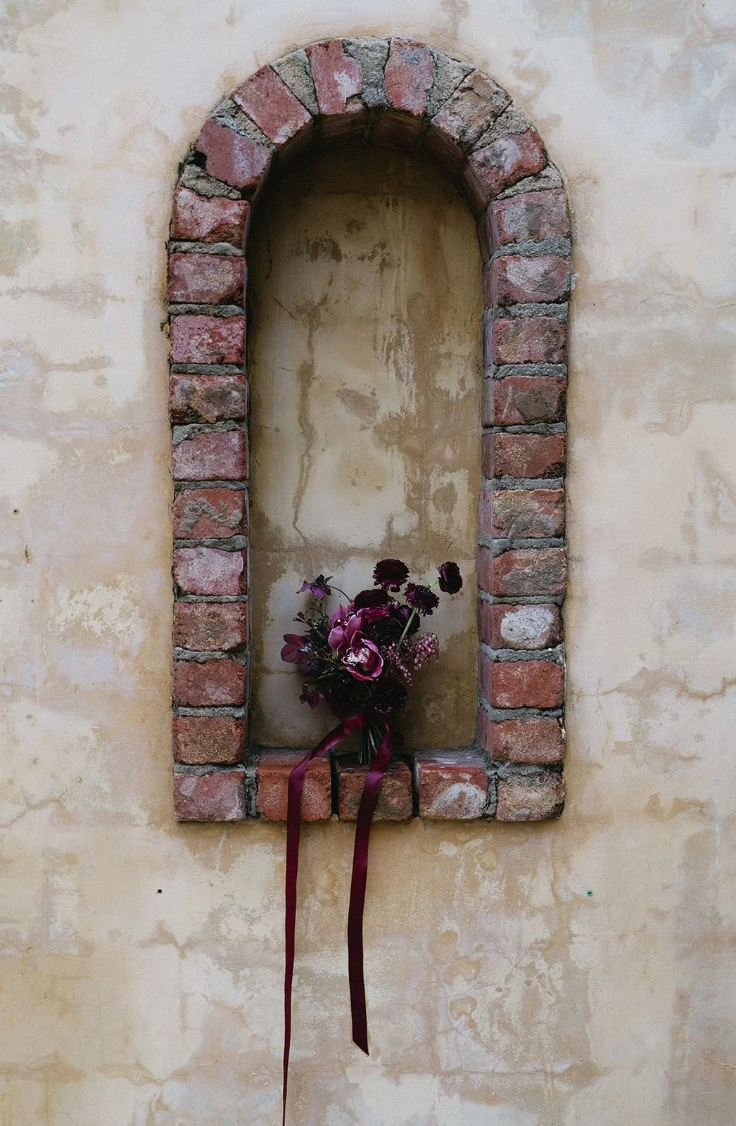A wall niche framed with worn red bricks, with a bouquet of dark purple and pink flowers resting inside, on a beige, textured wall.