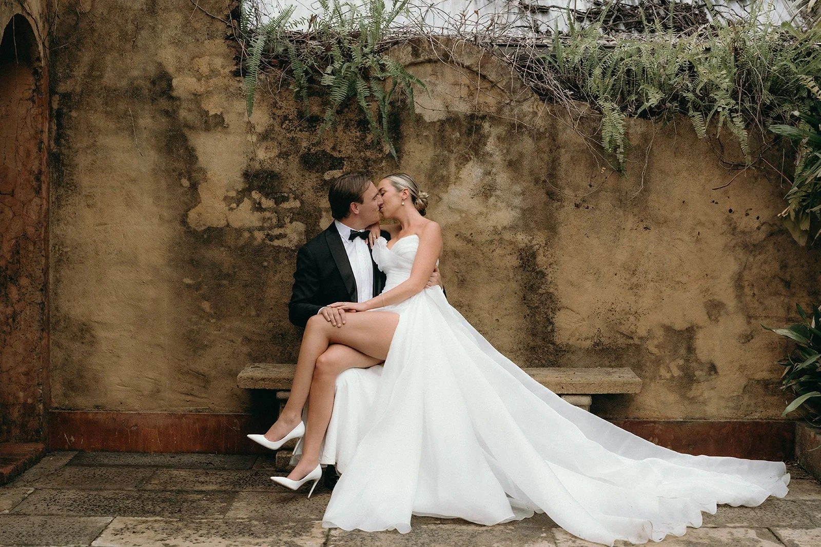 A bride in a white wedding dress and high heels sitting on a groom's lap, kissing, with a rustic wall background and greenery overhead.