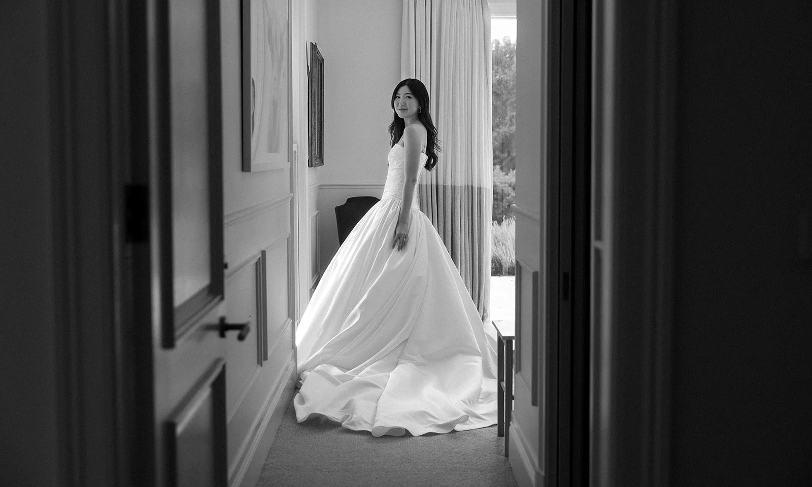 A woman in a wedding dress stands in a sunlit room, seen through a doorway, with curtains and framed pictures on the wall.