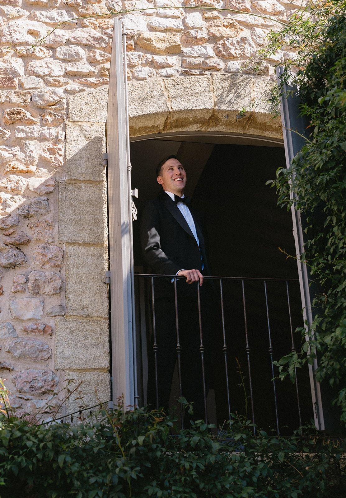 A man in a tuxedo standing on a small balcony, looking out and smiling, with a stone wall and greenery around him.