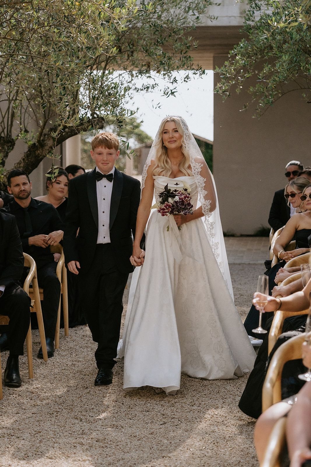 A bride in a white wedding dress and lace veil holding a bouquet of flowers walks down the aisle hand in hand with a young boy in a black tuxedo. Guests are seated on both sides, watching the ceremony outdoors under trees.