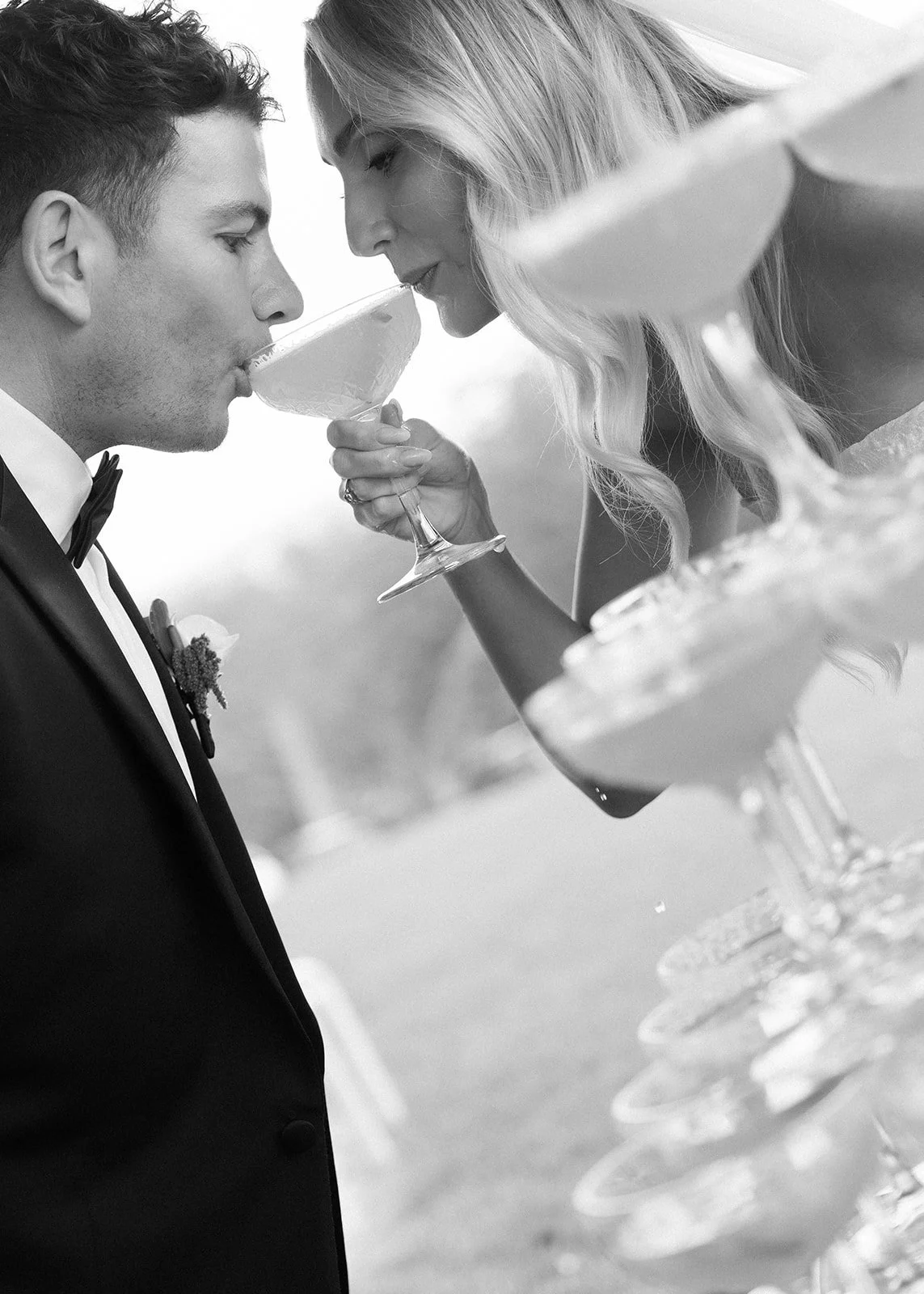 A black and white photo of a man and woman at a wedding, leaning in close with noses touching over a cocktail glass, with the woman holding the glass for the man to drink from.