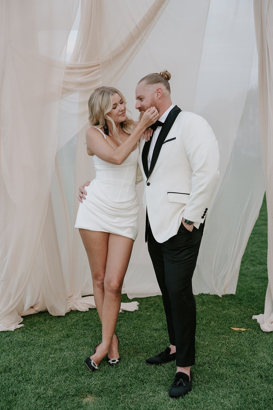 A woman and a man in formal attire sharing an intimate moment outdoors with draped fabrics in the background.