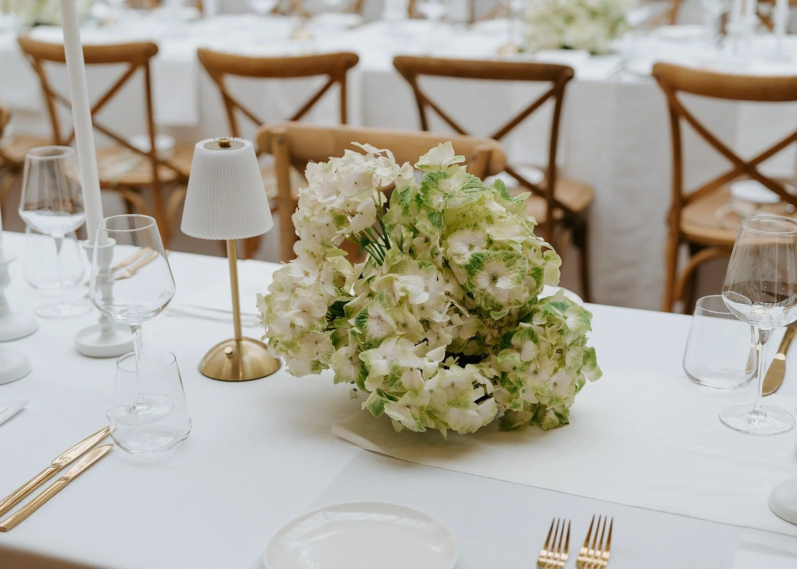 Elegant dining table with a white tablecloth, large floral centerpiece with white and green flowers, surrounded by glassware, gold utensils, a small white lamp, and wooden chairs in the background.