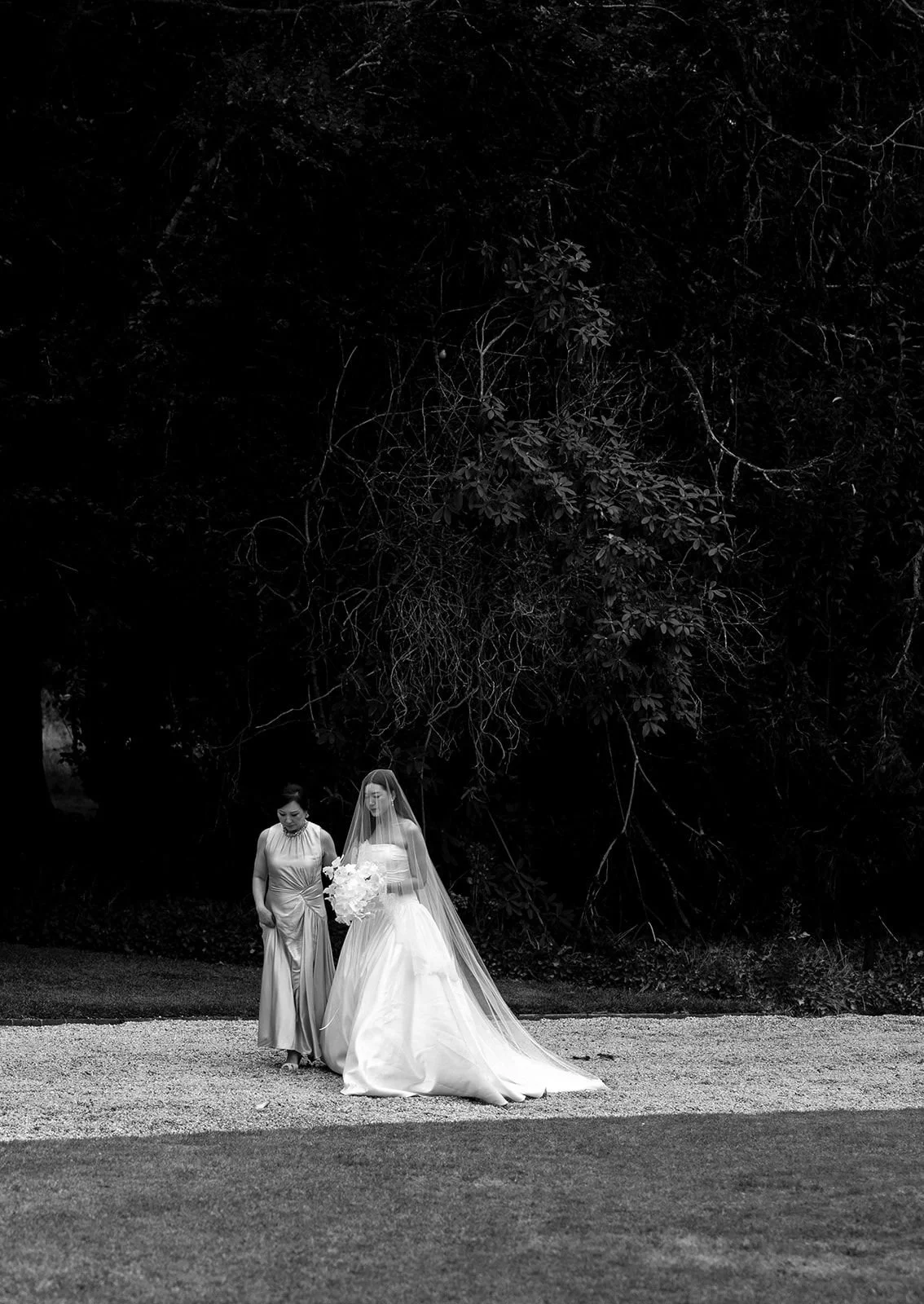 A bride in a wedding dress and veil holding a bouquet walking beside a woman in an elegant gown, outdoors near trees.