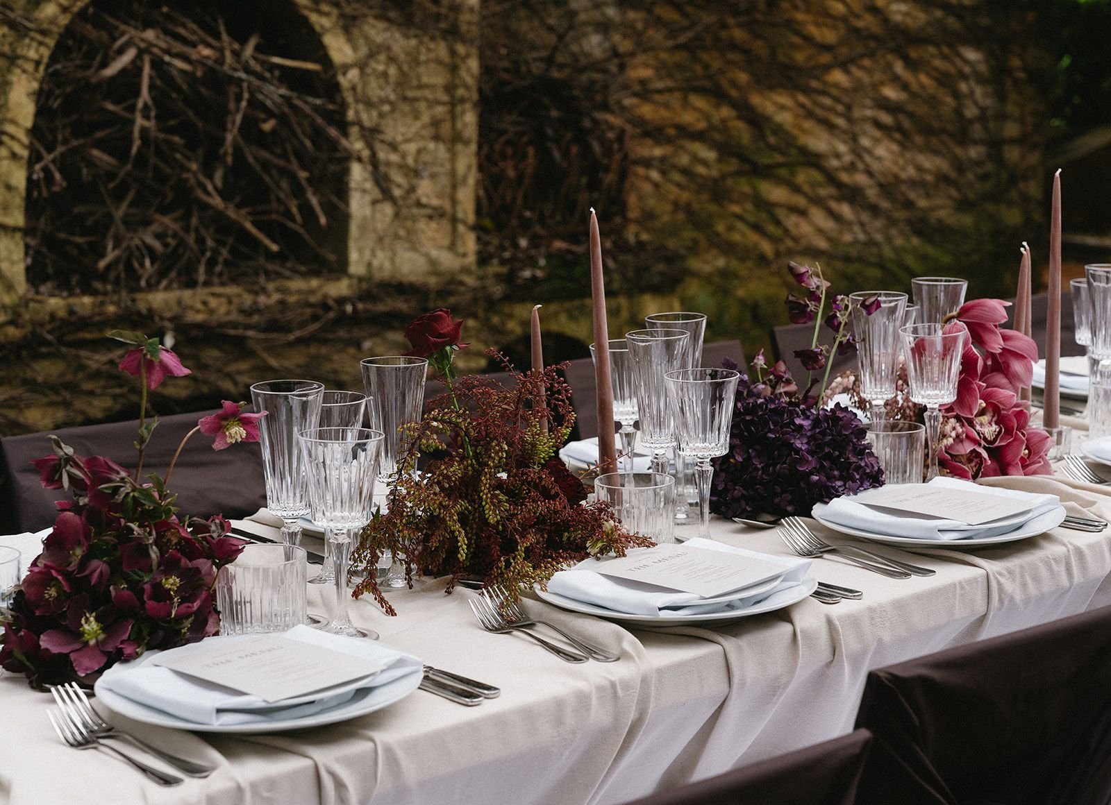 Elegant dining table set with white tablecloth, floral centerpieces, pink and purple flowers, crystal glassware, white plates, silver cutlery, pale pink taper candles, and black chairs, in front of a rustic stone wall with branches.