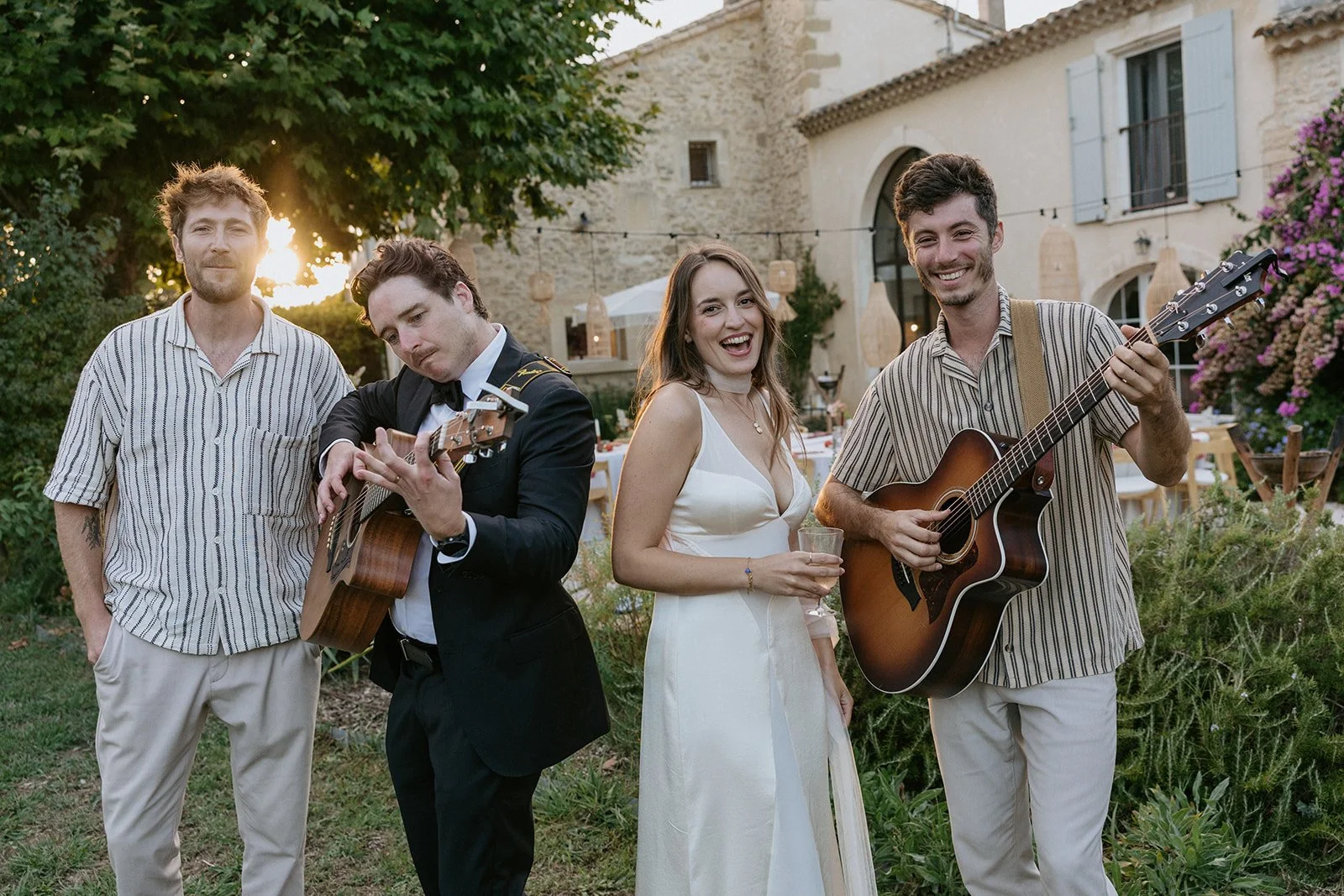 Group of four people outdoors at sunset, two men playing guitars, a woman in a white dress holding a drink, all smiling, in a garden with greenery and an old stone building in the background.