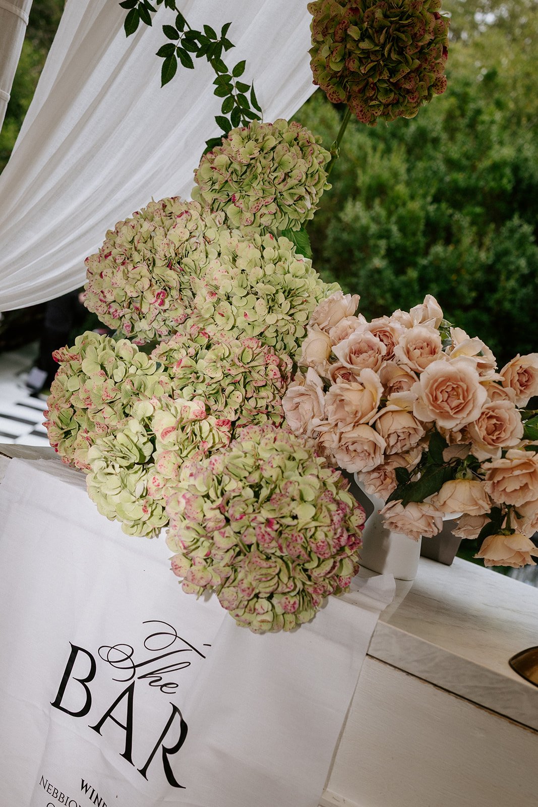 Bouquet of light pink roses and green hydrangeas with pink accents, in a white vase, on a white table with a sign that reads 'The Bar' in black text, set outdoors with greenery in the background.