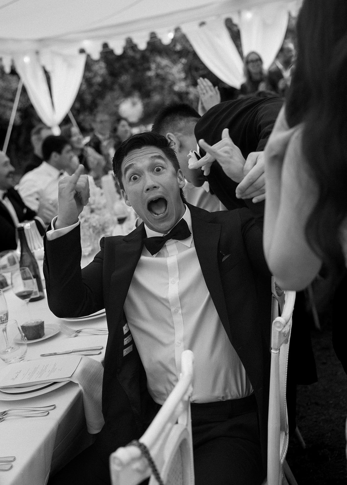 A man wearing a tuxedo and bowtie at a formal event, with a surprised or excited expression, mouth open, and pointing upwards with one finger. People sit at dressed tables in the background under a decorated tent.