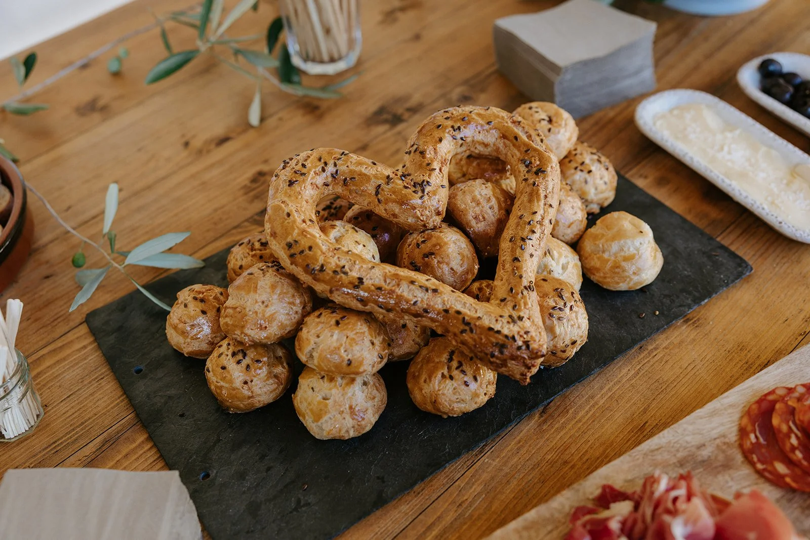 A black rectangular platter with small bread rolls topped with black sesame seeds, arranged around a heart-shaped pretzel on a wooden table.