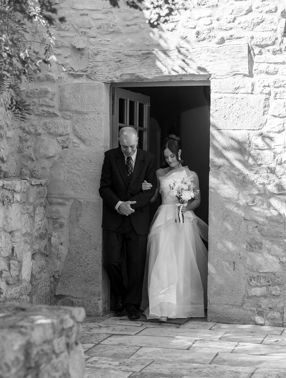 Black and white photo of a bride in a wedding dress holding a bouquet, walking with an older man in a suit through a stone doorway.
