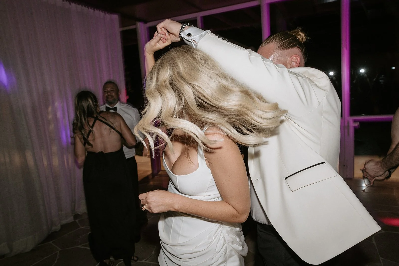 Couple dancing at a wedding reception, woman with blonde hair in a white dress, man in a white suit jacket, other guests dancing in the background.