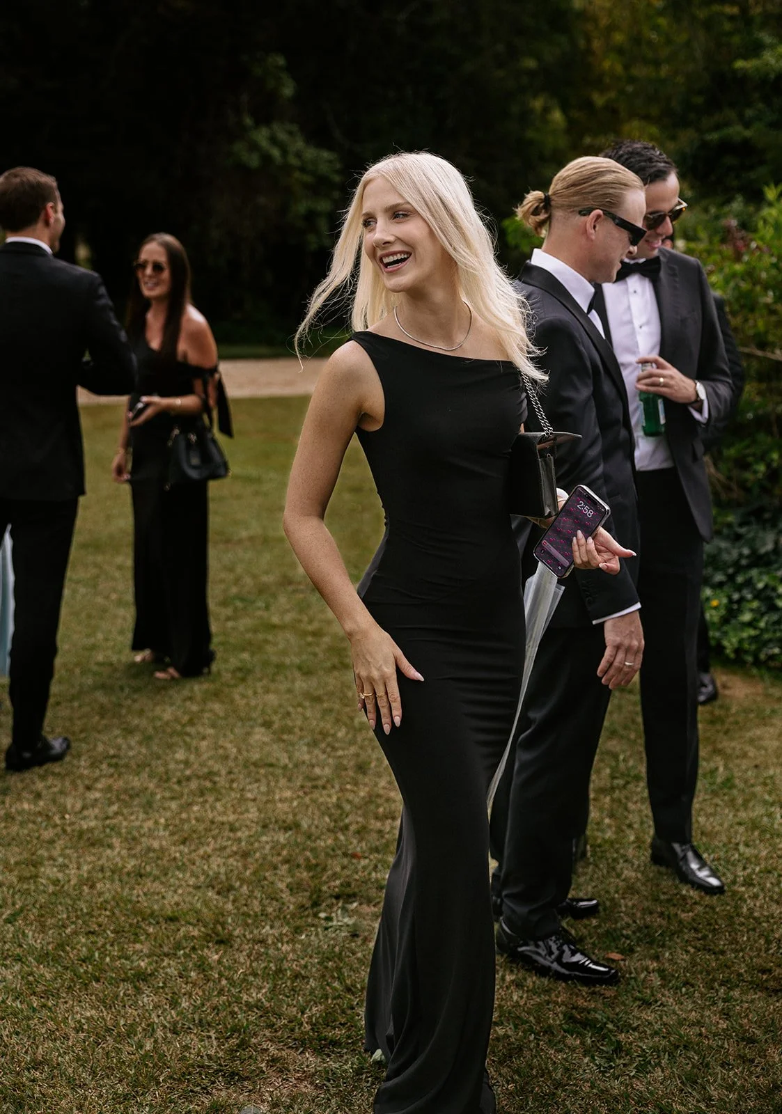 A group of people dressed in formal black attire, standing outdoors on grass, at an event or gathering, with trees in the background. The focus is on a smiling blonde woman in a black dress holding a phone and an umbrella.