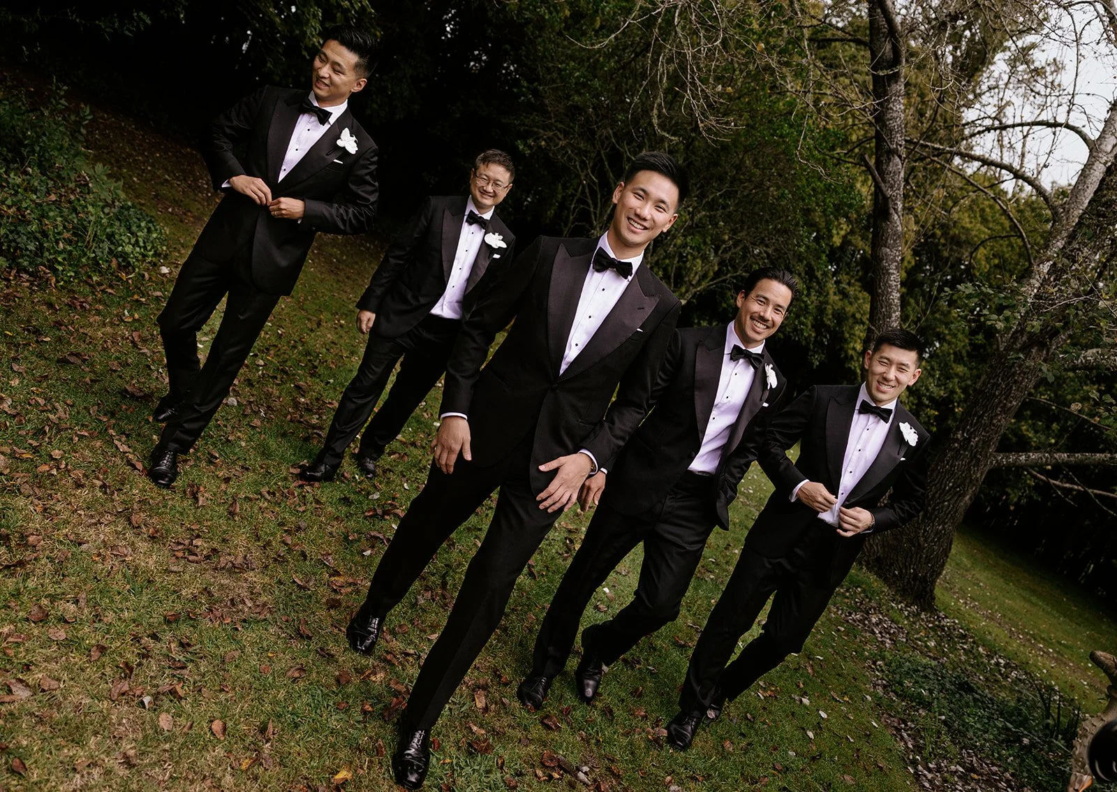 A group of five men dressed in tuxedos with white shirts and black bow ties, standing outdoors on grass with trees in the background, smiling and posing for the photo.