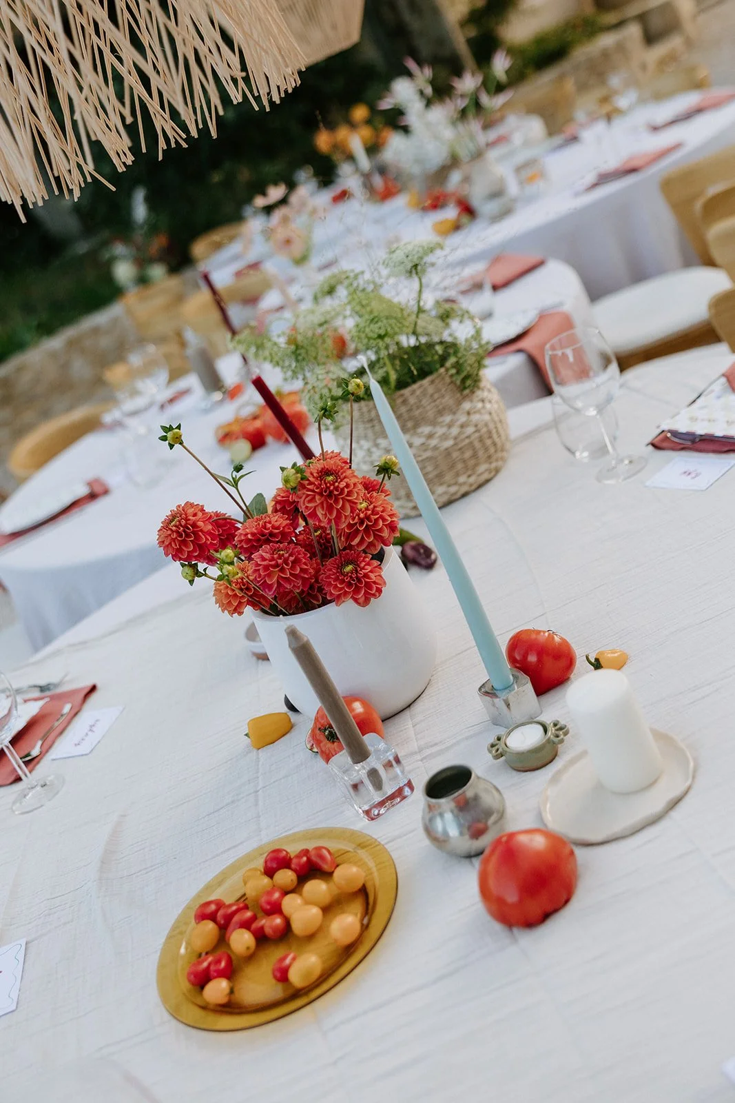 A table set for an outdoor event with a centerpiece of red and orange flowers, candles, and assorted fruits, including tomatoes and small yellow fruits.