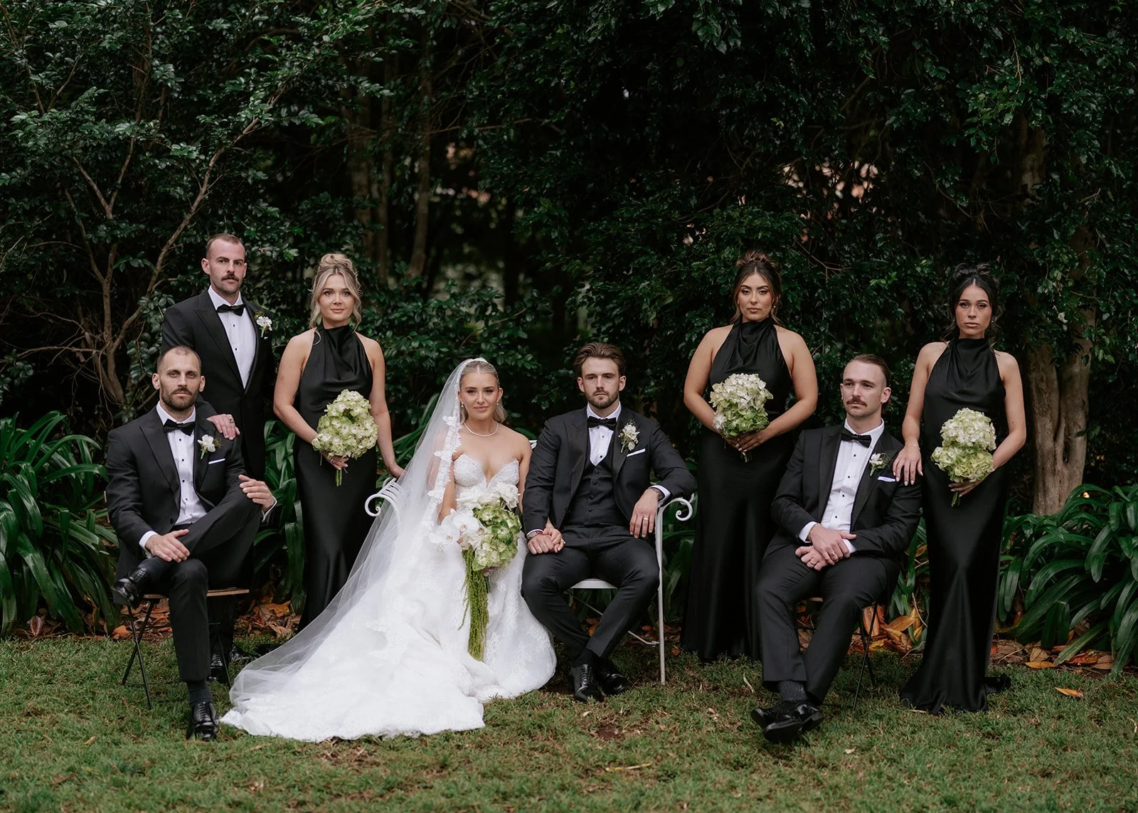 Group of nine people dressed in formal attire, posing outdoors for a wedding photo with lush greenery in the background. The bride wears a white gown and veil, holding a bouquet, seated next to the groom in a black tuxedo. Four bridesmaids in black d