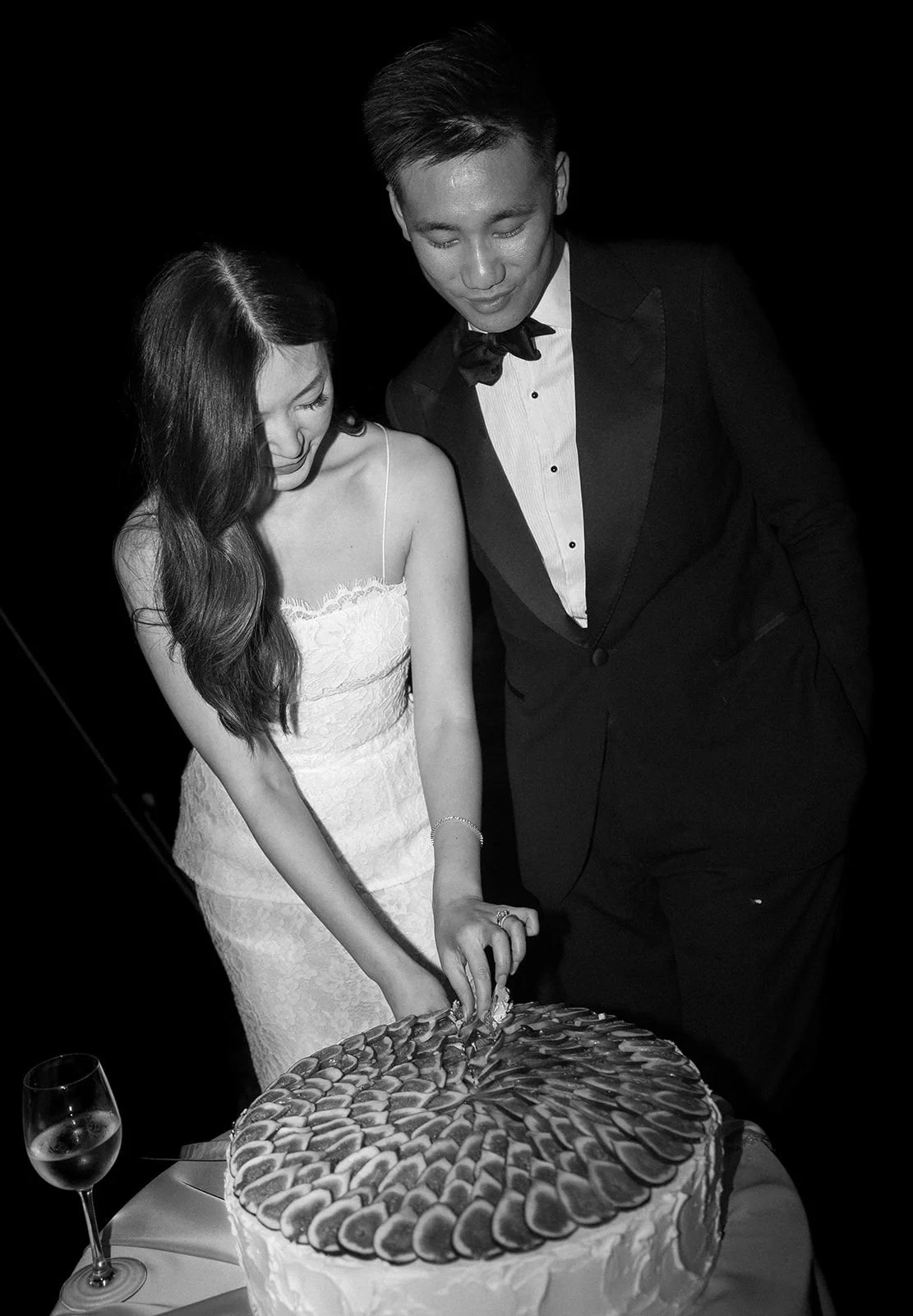 A black-and-white photo of a bride and groom cutting a wedding cake together. The bride is wearing a white strapless dress, and the groom is dressed in a tuxedo with a bow tie. A glass of wine is on the table.