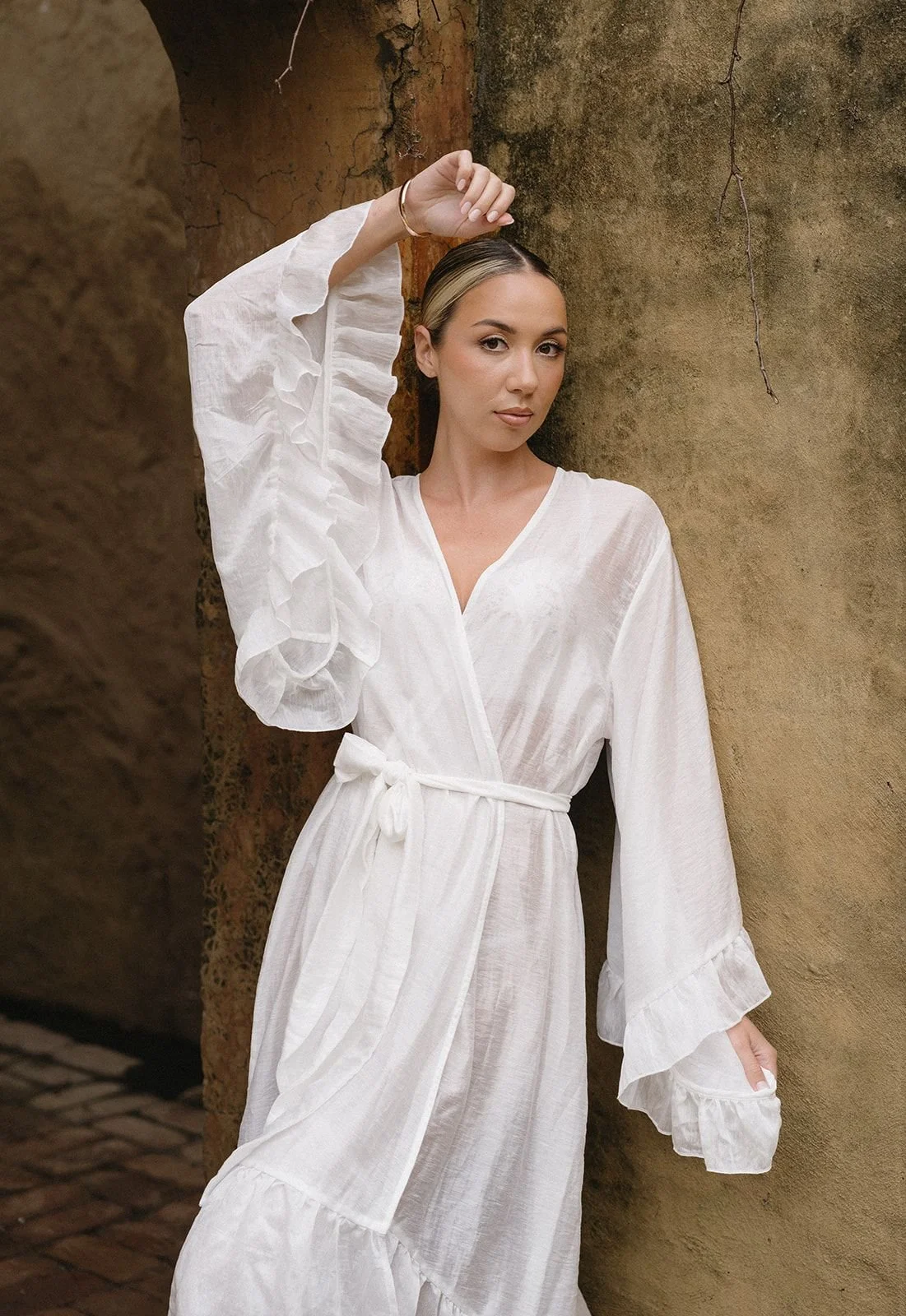 A woman with blonde hair tied back, wearing a white, long-sleeved dress with ruffled cuffs and a tie at the waist, standing against a rustic, textured wall, with one arm raised above her head.