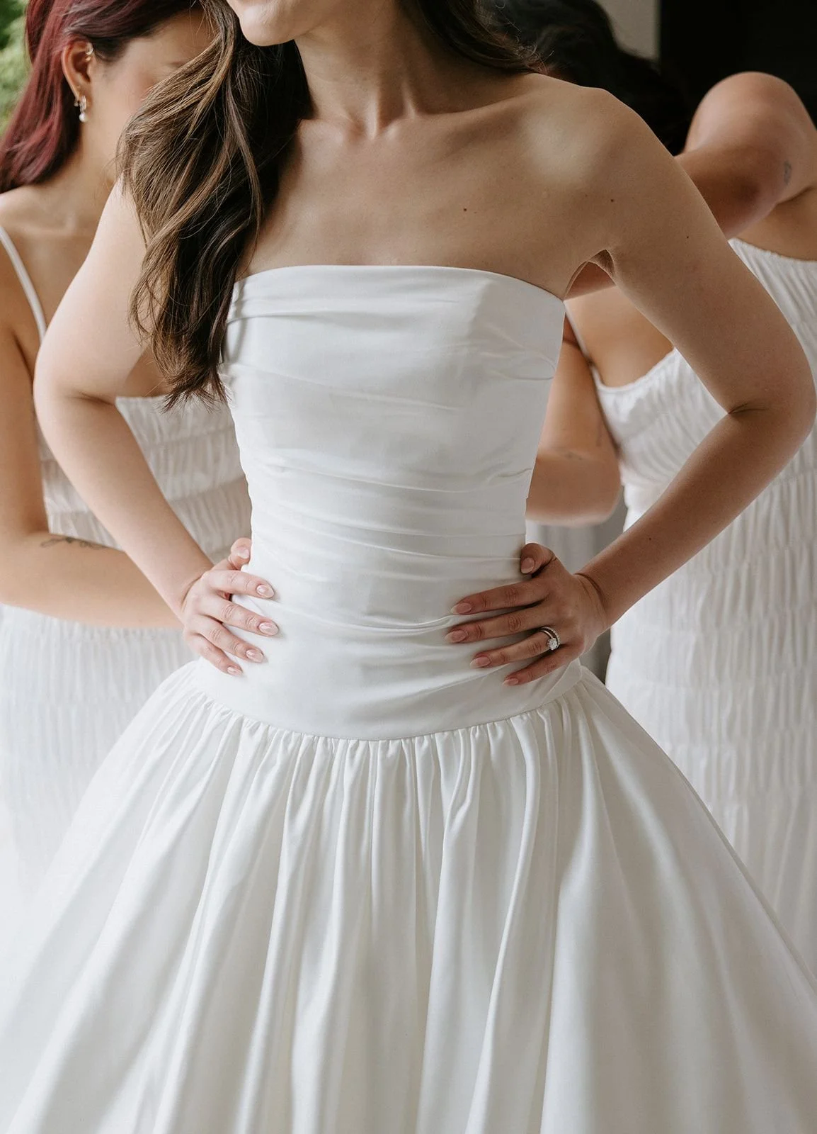 A bride trying on a white strapless wedding gown with pleated bodice and full skirt, surrounded by bridesmaids in matching dresses.