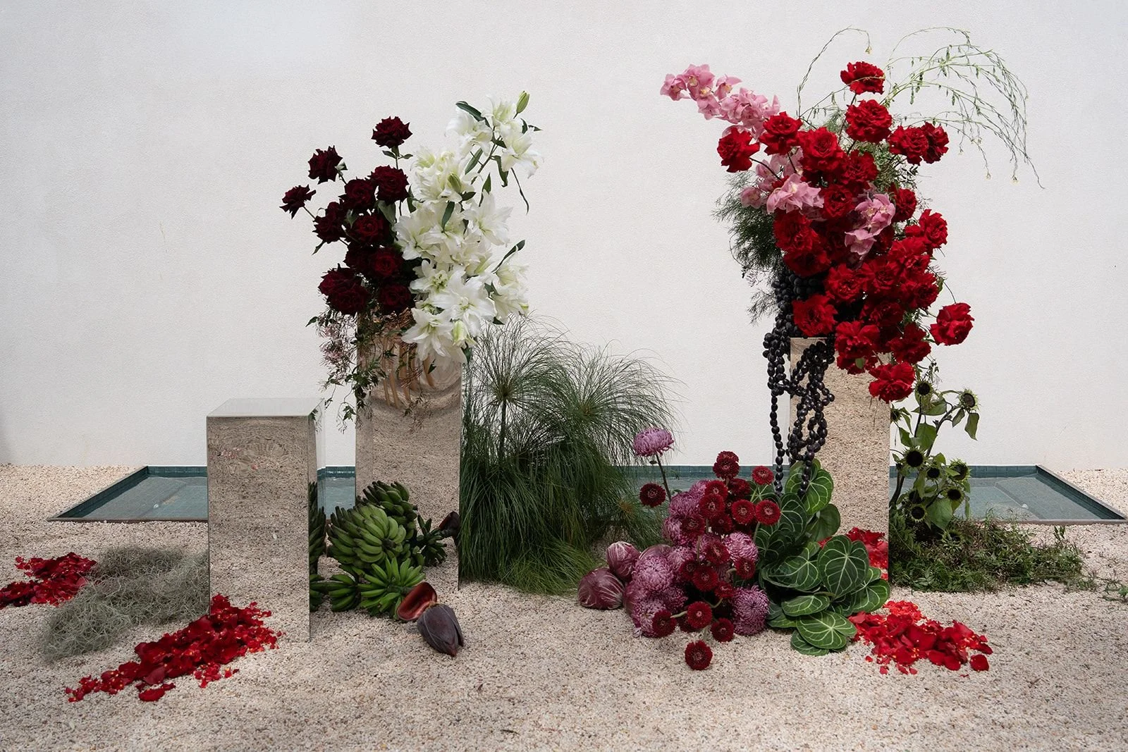 Arrangement of various flowers including red, white, and pink blooms, placed on a sandy surface with mirrored blocks and some greenery.