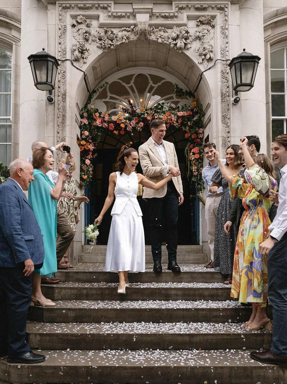 A joyful couple leaving a wedding venue, holding hands and smiling, surrounded by friends and family celebrating on the steps decorated with flower arrangements and confetti.