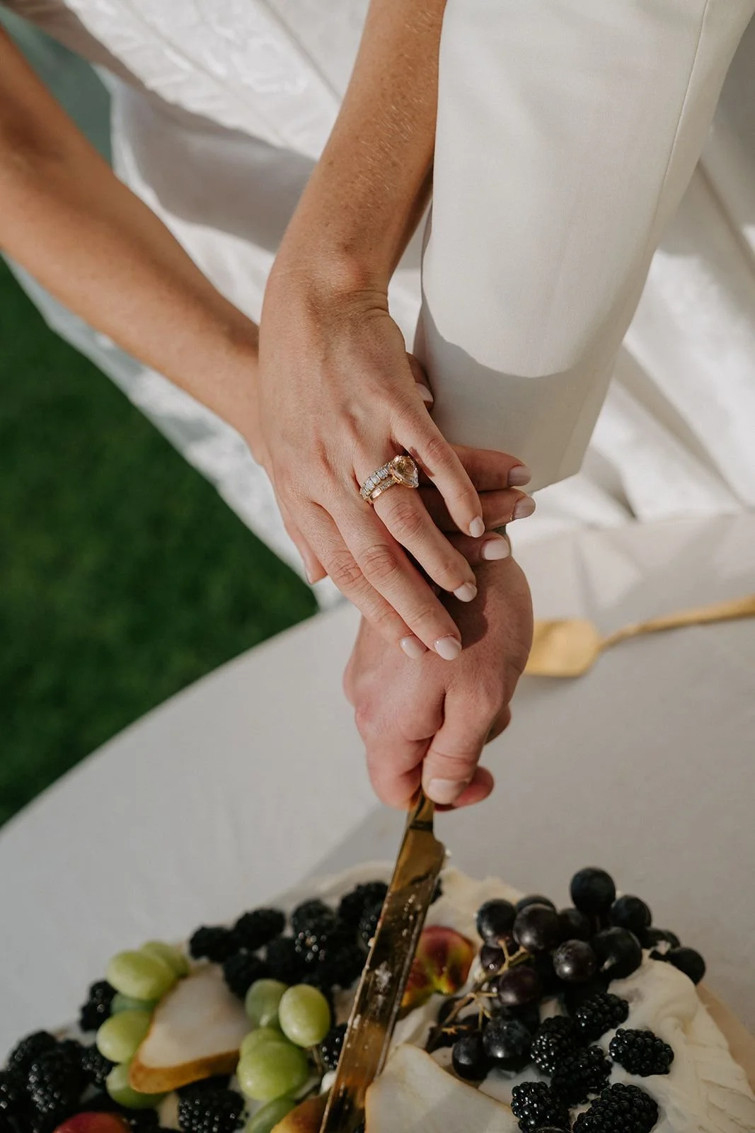 A bride wearing a wedding ring is cutting a wedding cake decorated with blackberries, black grapes, green grapes, and pear slices.